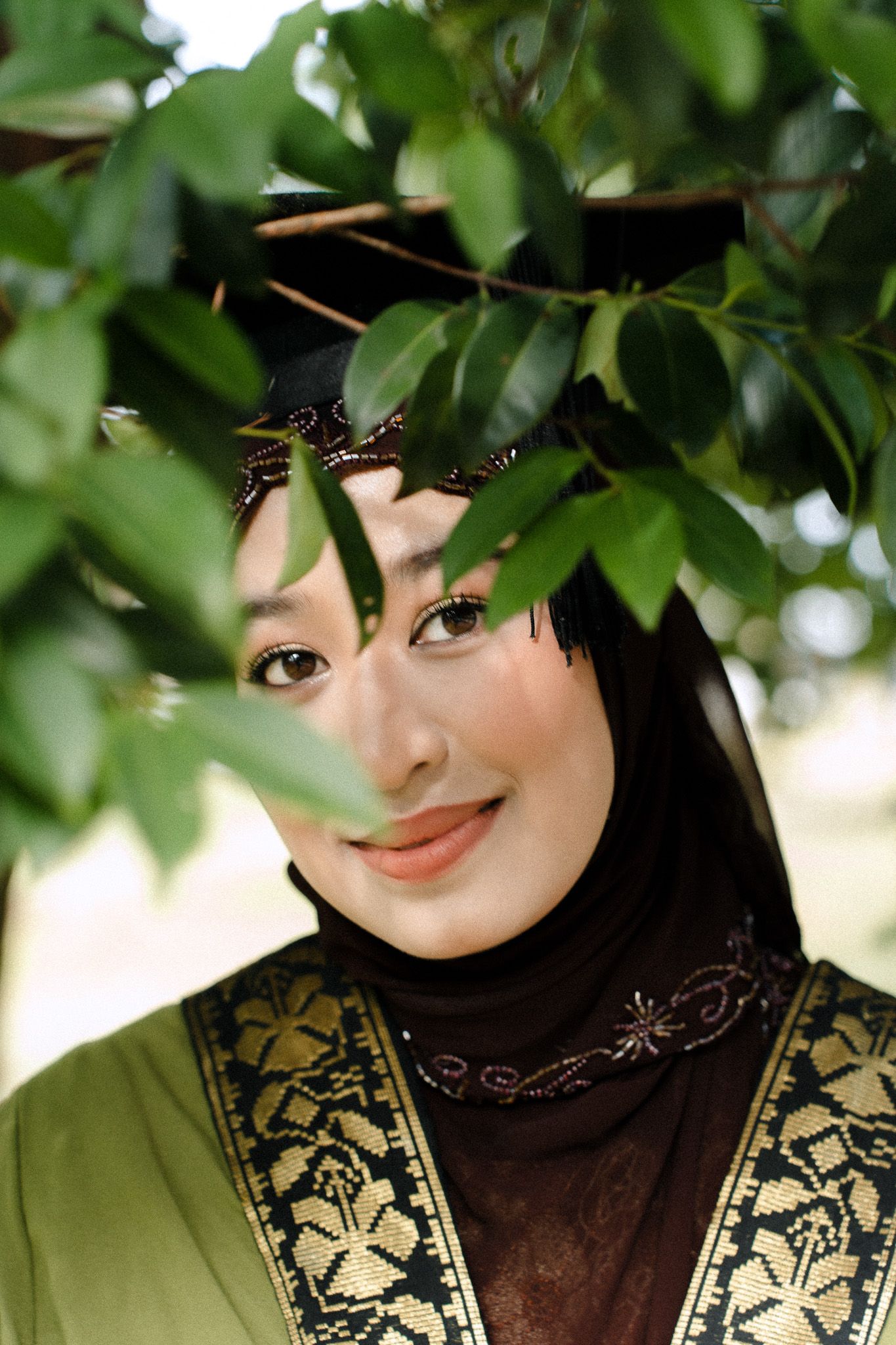 A smiling woman in a hijab and traditional green embroidered dress poses among lush green tree branches outdoors.