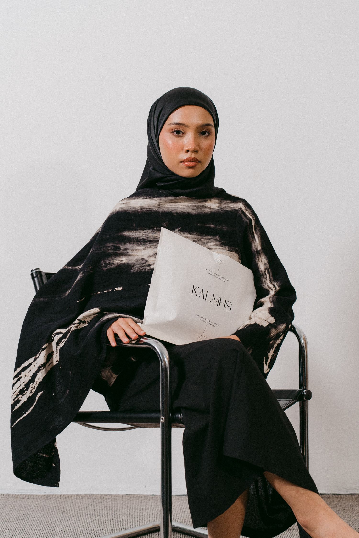 Model in black and white outfit with hijab holding shopping bag while seated in chair.