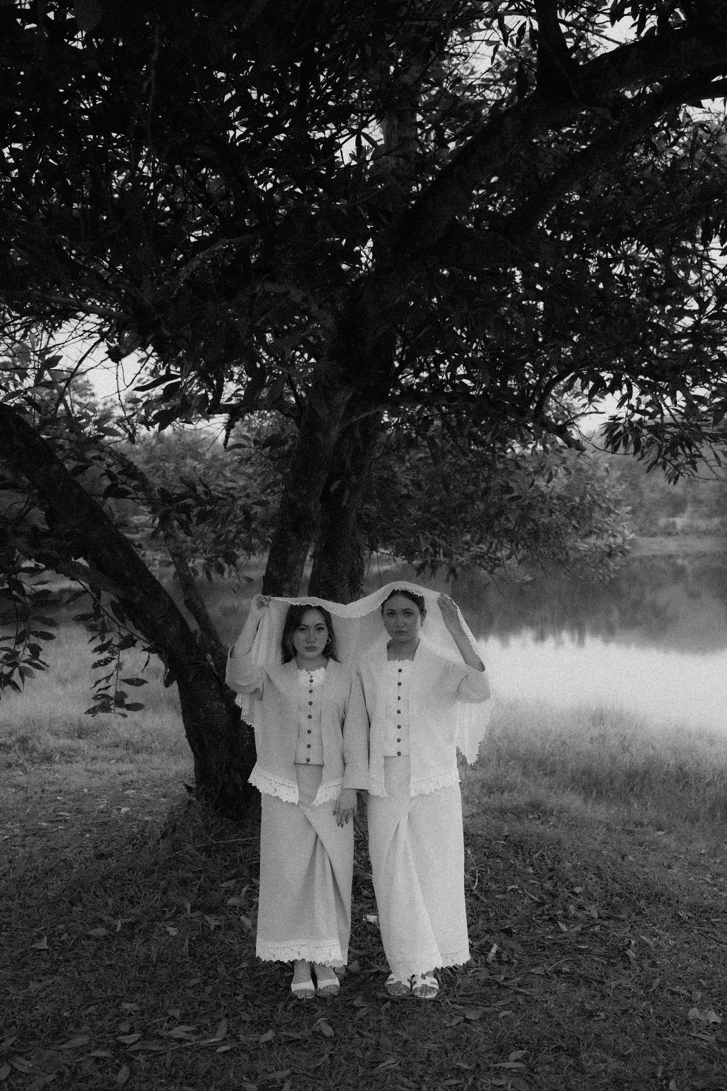 A vintage black and white photo of two figures in white clothing standing under a tree beside a misty lake.