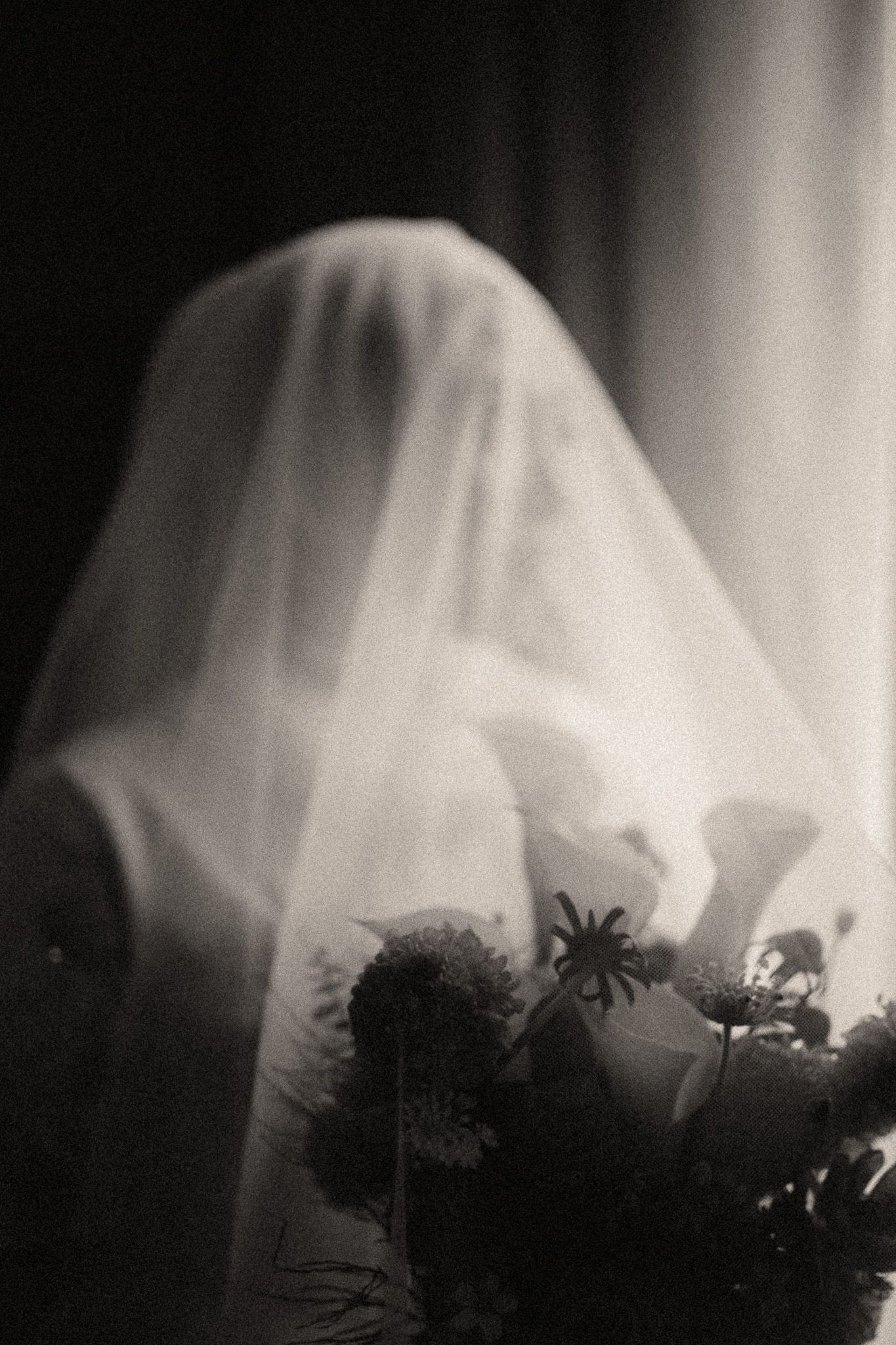 Eerie black and white photo of a ghost-like figure draped in white veil holding a bouquet of flowers.