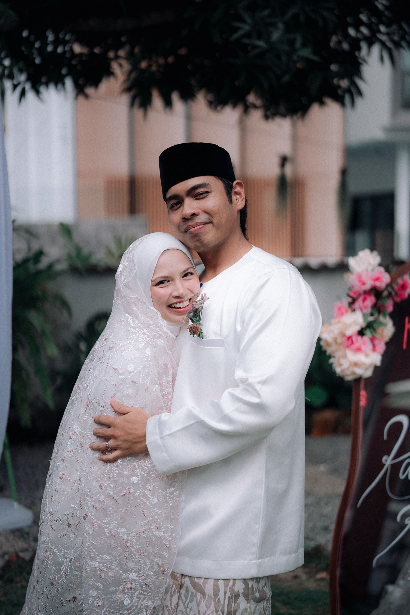 Muslim couple in white traditional attire embracing outdoors, woman in hijab and lace dress, man in baju melayu and songkok.