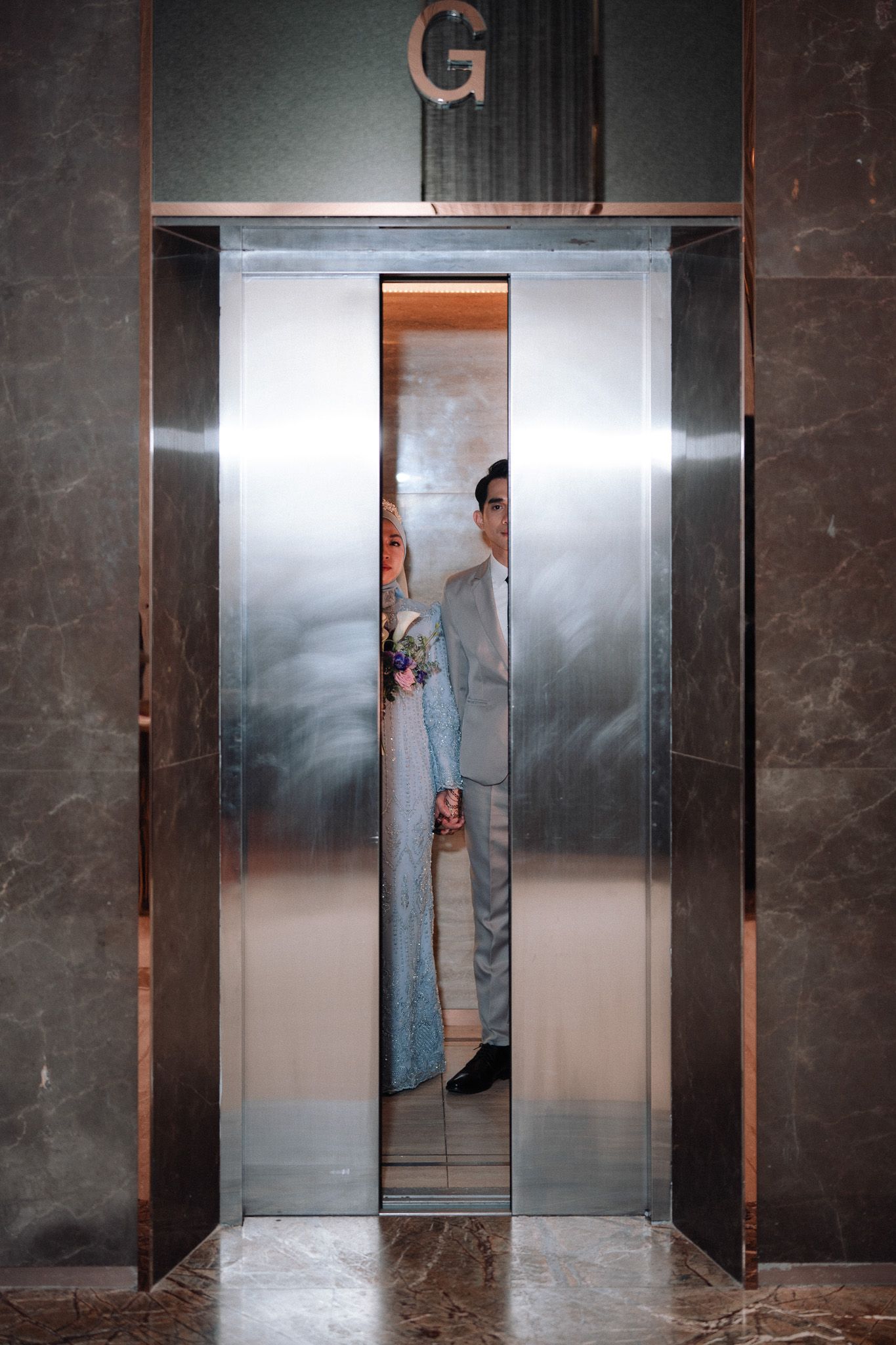 A bride and groom stand together in an elegant elevator with gold trim, the bride holding a bouquet.
