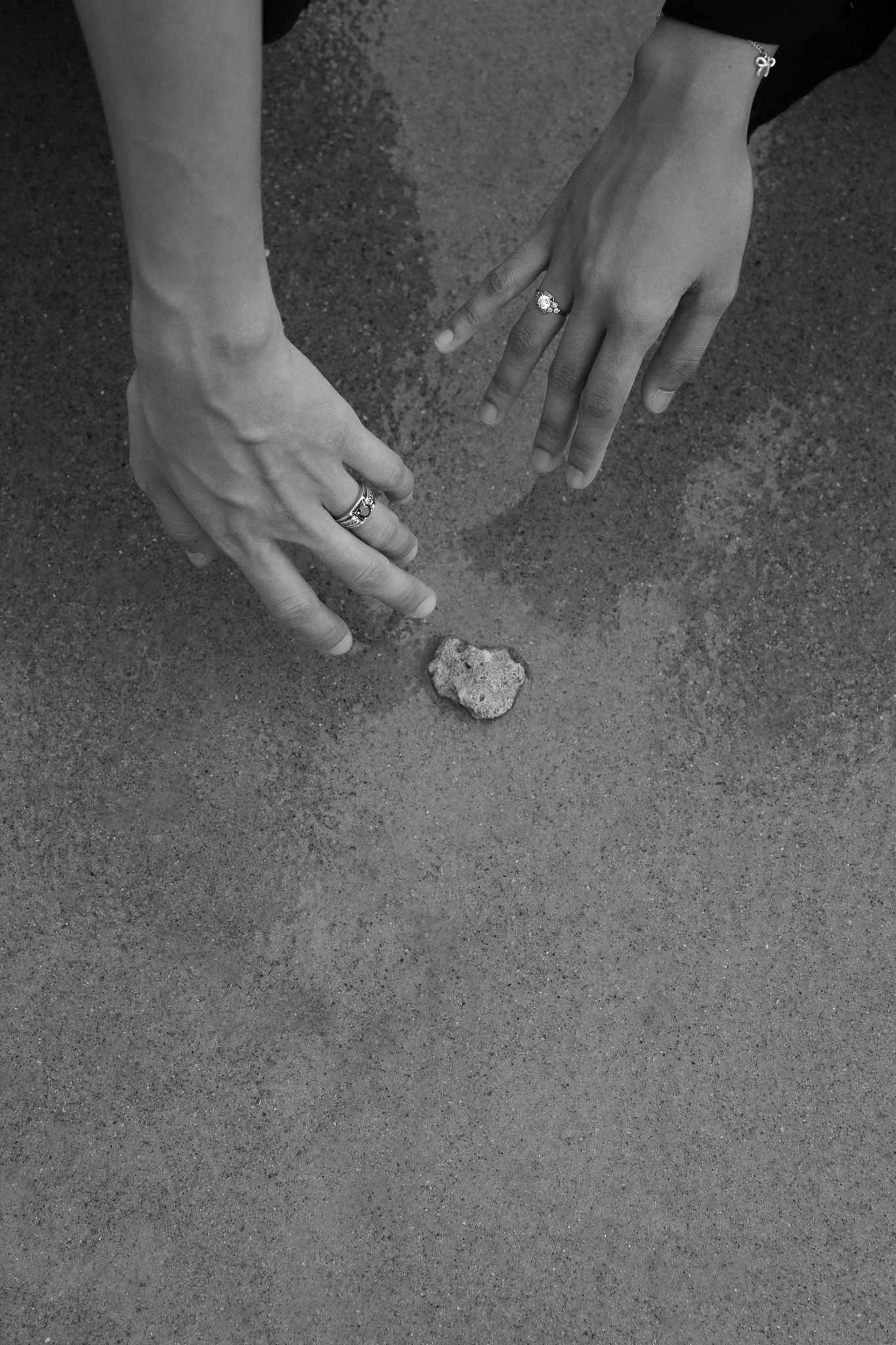 Black and white photo of two hands with rings reaching toward a small rock on a textured concrete surface.