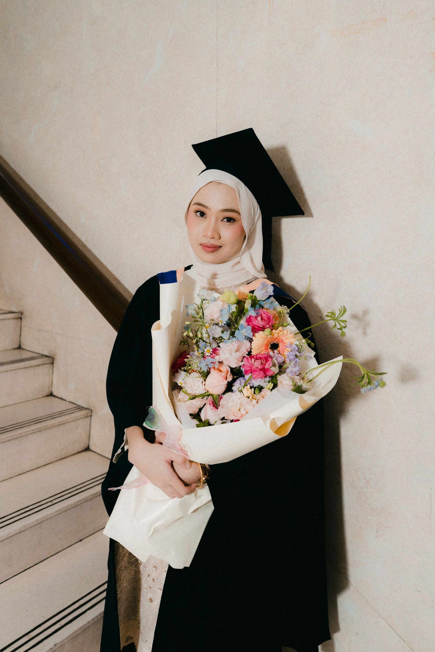 Graduate in black cap and gown holding a vibrant bouquet of pink and white flowers on white stairs.