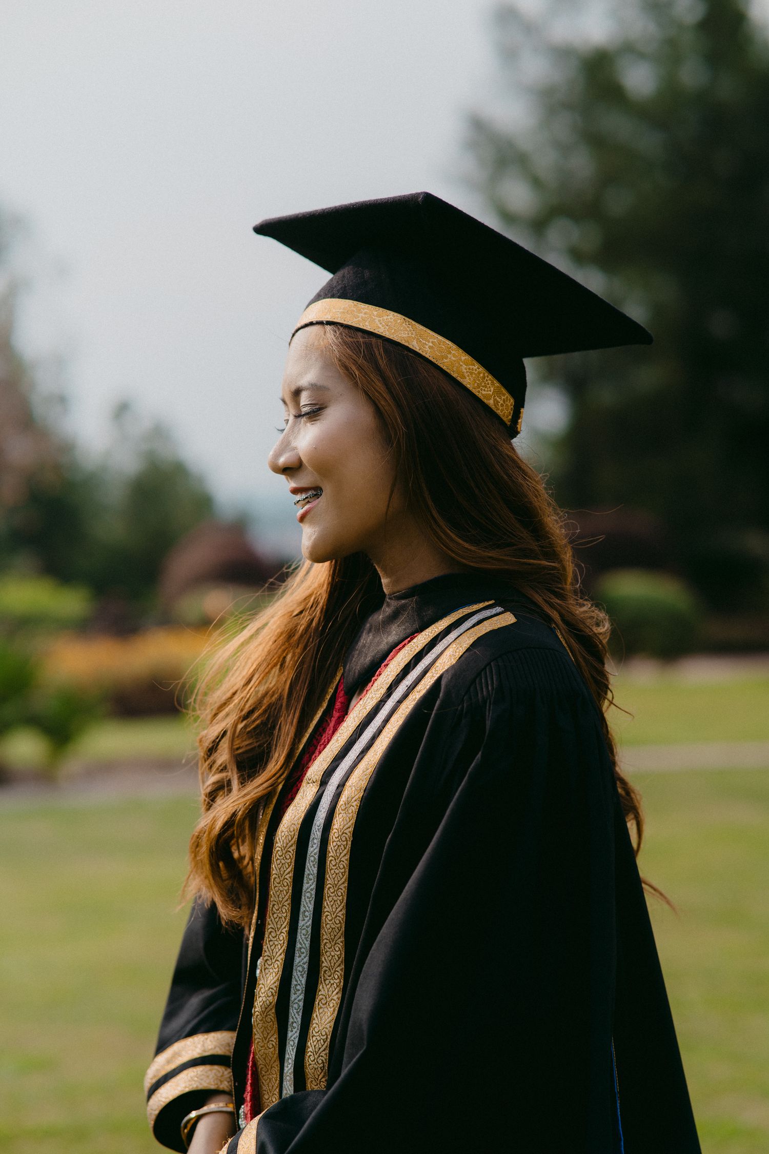 Graduate in black cap and gown with gold trim smiles while posing outdoors.