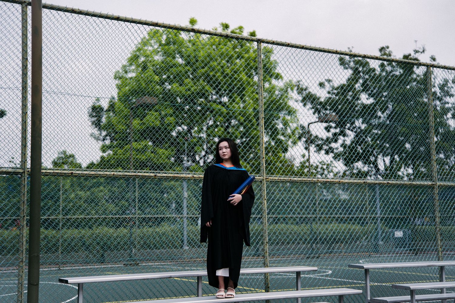 Graduate in black gown standing on bleachers at outdoor sports facility.