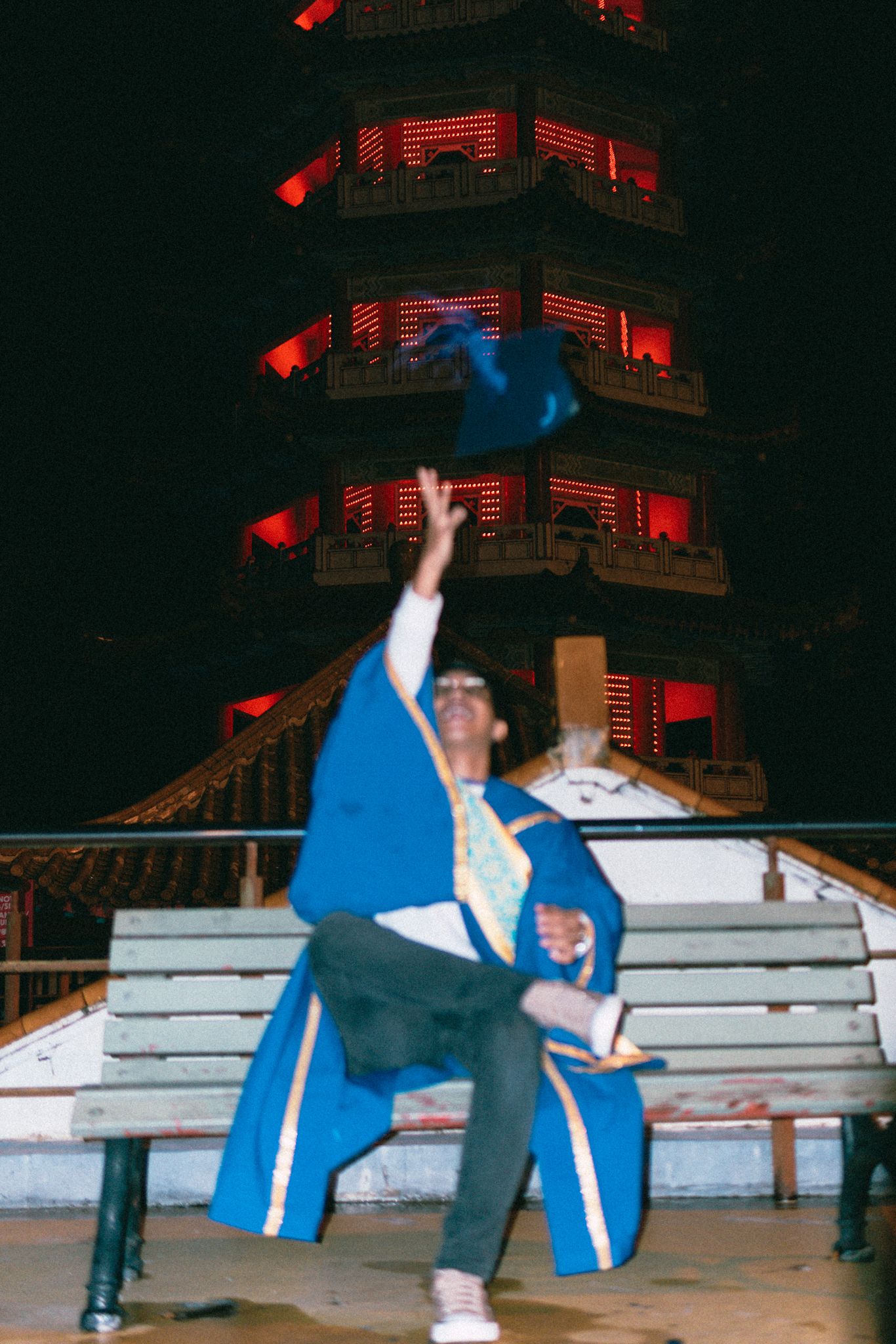 Graduate in blue gown striking dramatic pose in front of lit pagoda at night.