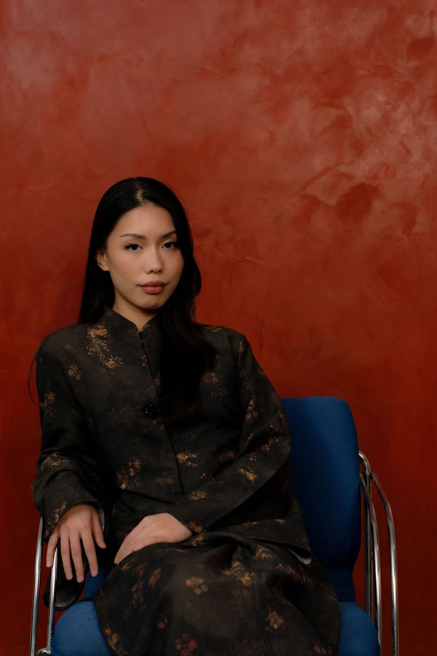 Person in traditional black floral Chinese dress sits elegantly on blue chair against red textured wall.