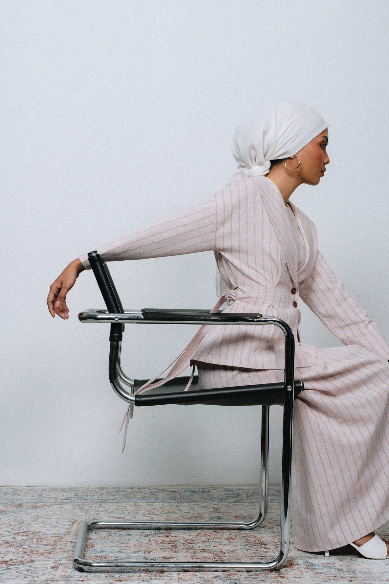 Woman in striped suit and white head wrap seated on modern chrome chair against light gray wall.
