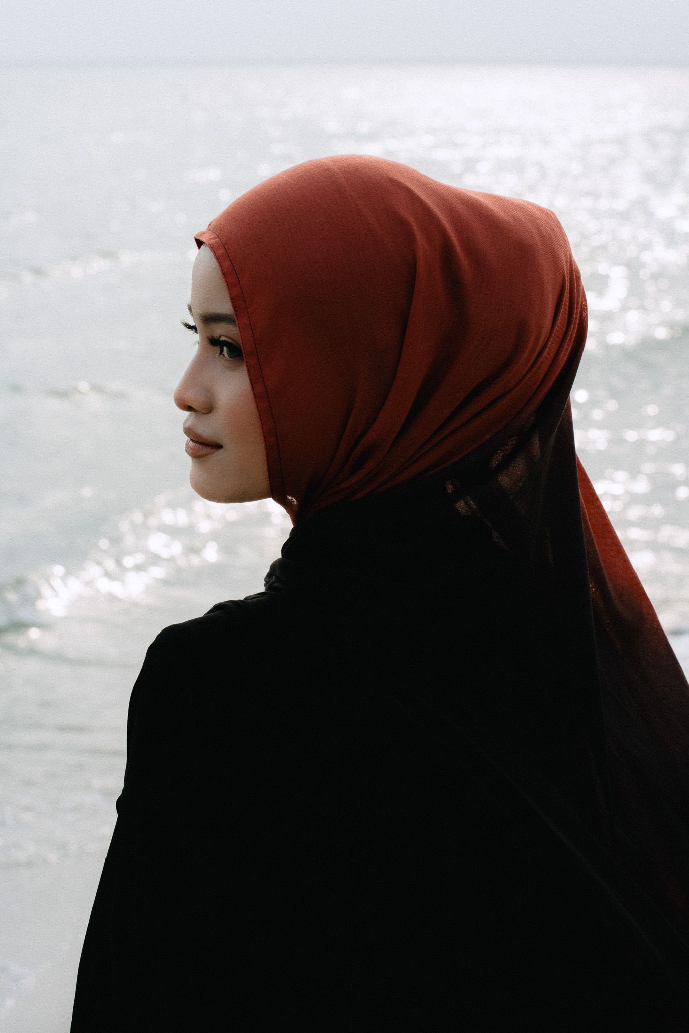 Young woman wearing a rust-colored hijab and black abaya standing by the ocean, gazing thoughtfully into the distance.