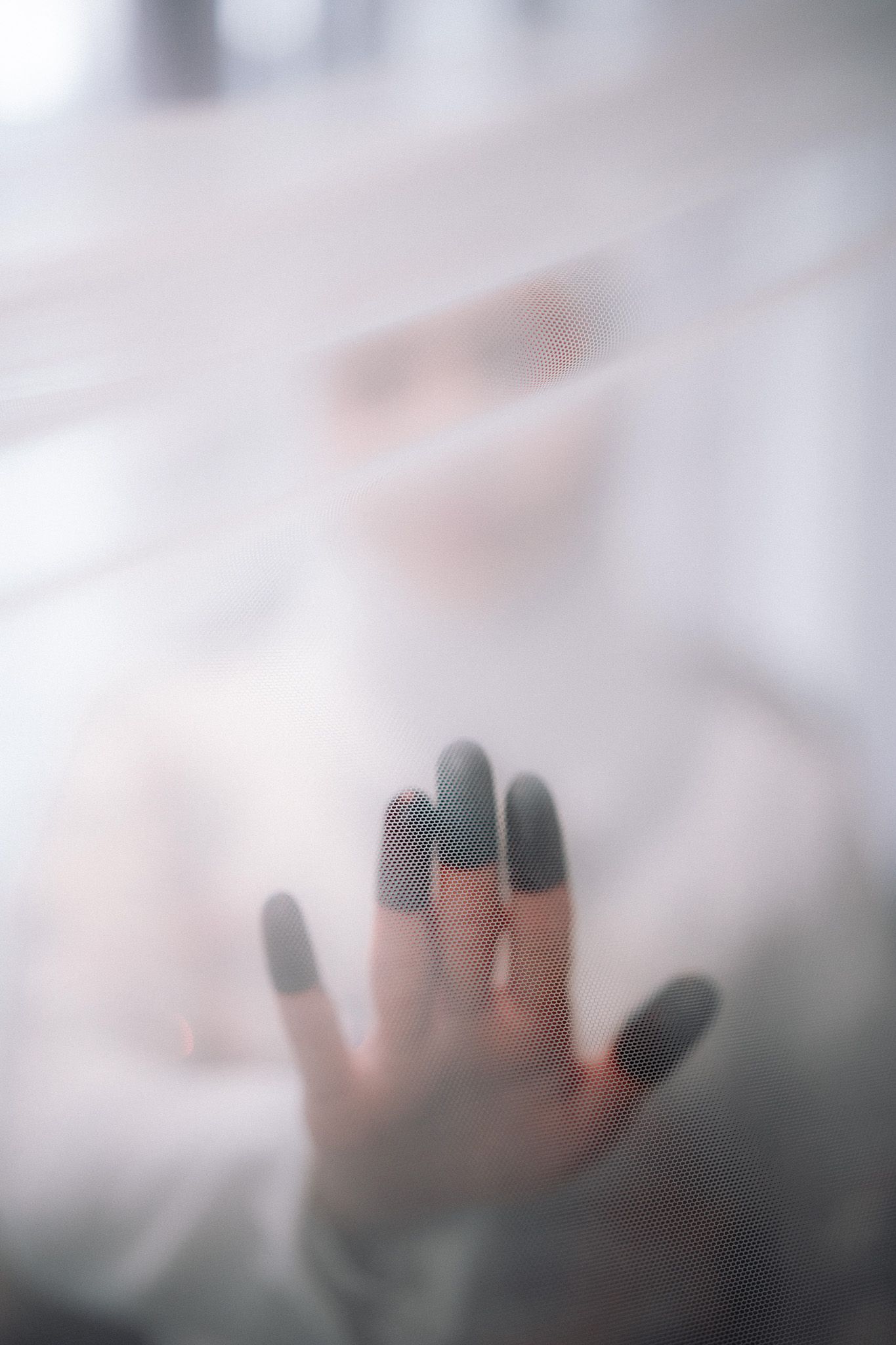 A blurred hand with dark fishnet glove fingertips pressed against sheer white fabric with a face visible behind it.