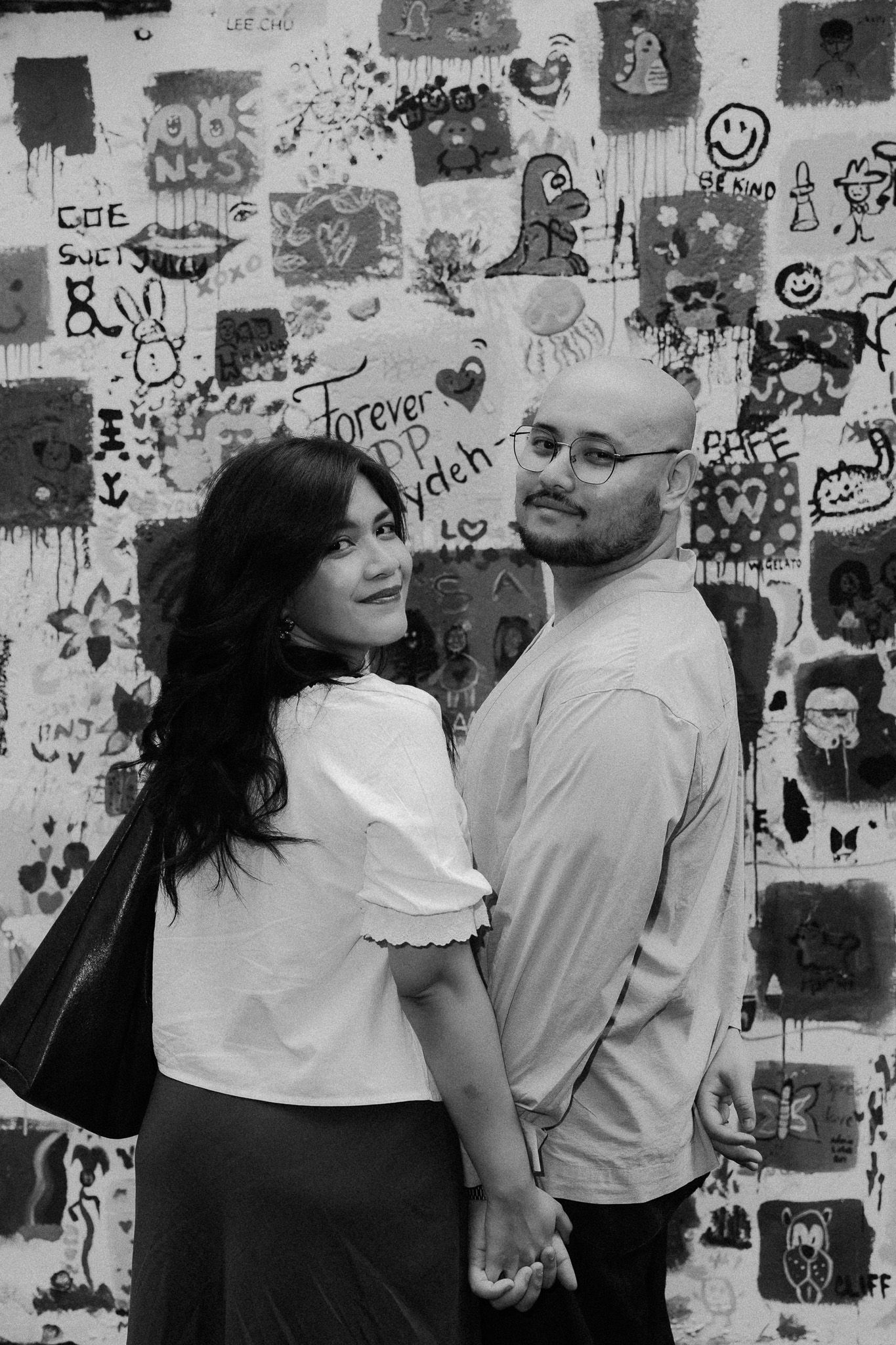 Black and white photo of a couple holding hands in front of a graffiti wall with doodles and text.