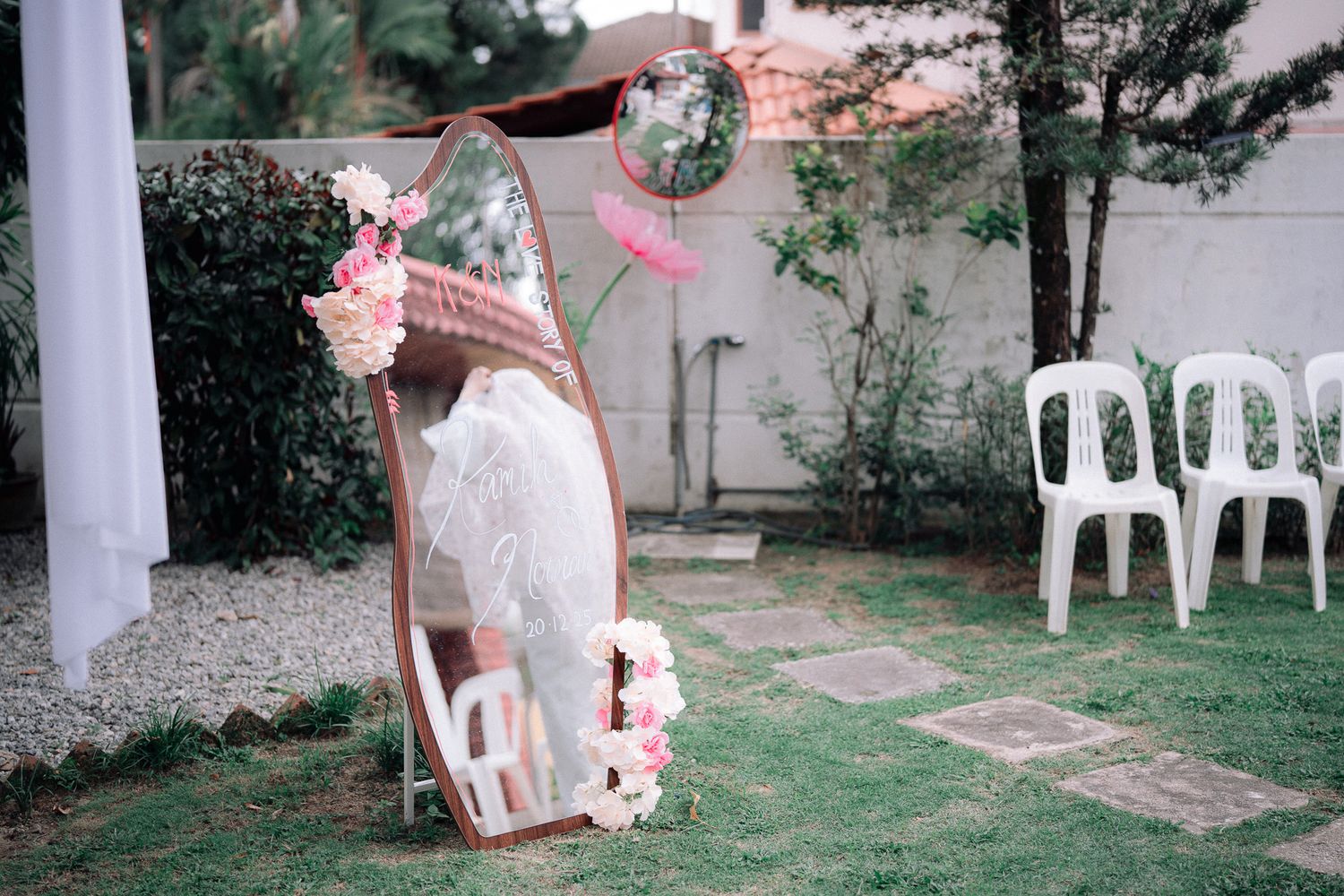 Elegant outdoor wedding setup with floral mirror, white chairs on green lawn, and pink flower decorations.