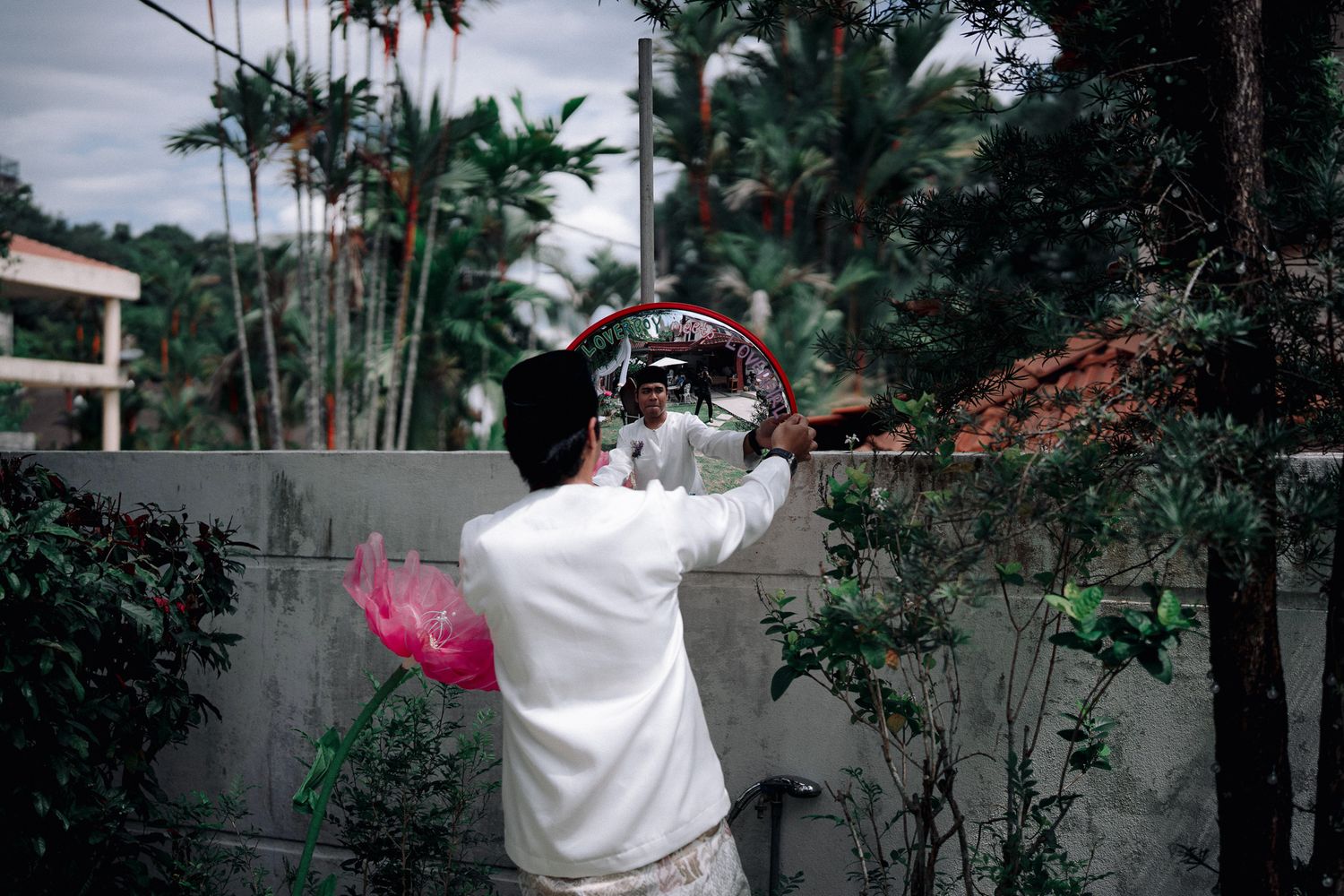 Person in white traditional attire holding a decorative circular object outdoors among lush tropical trees and greenery.