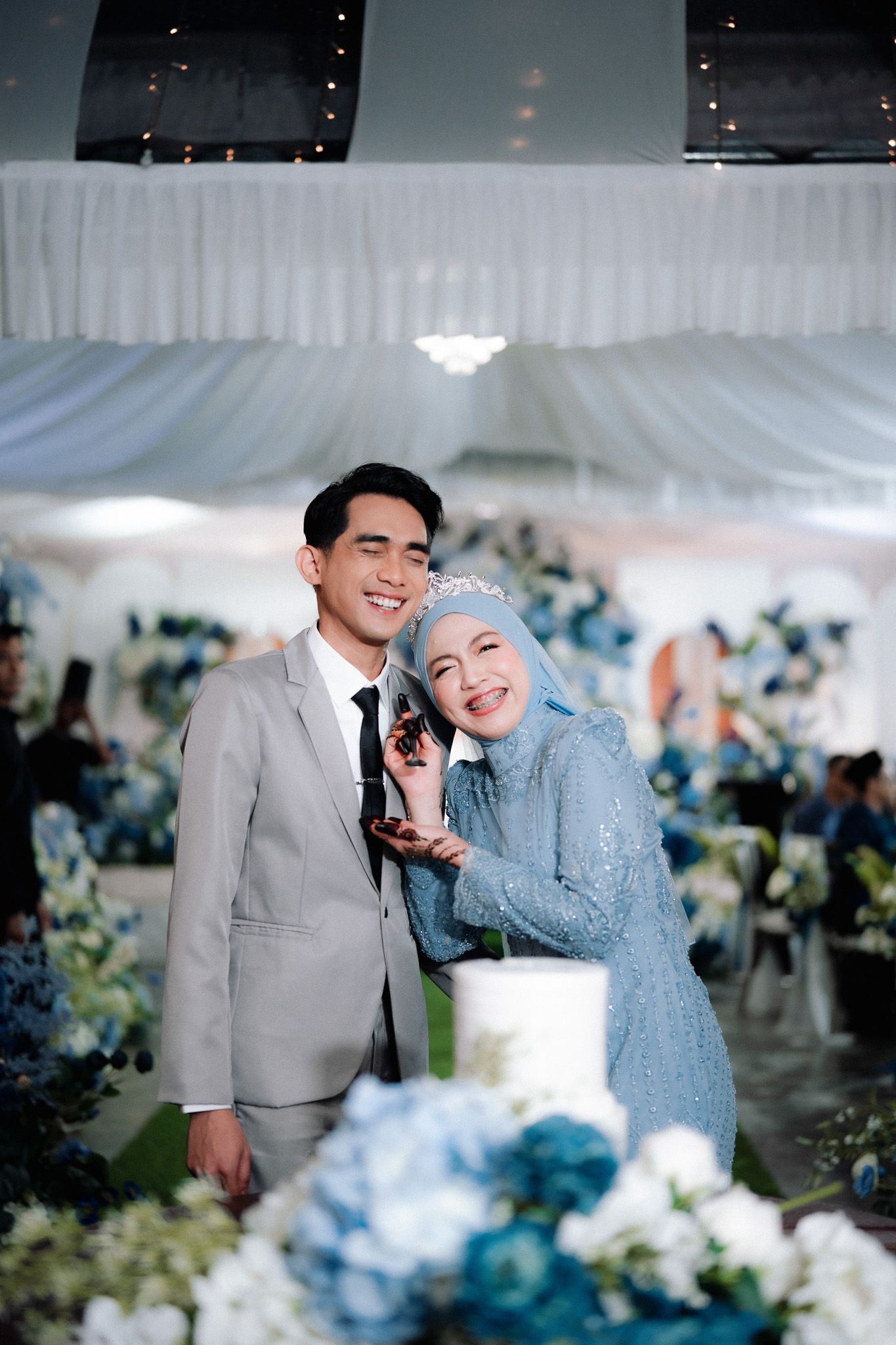 A smiling couple in elegant blue and gray wedding attire poses beside floral arrangements and a white cake at a reception.