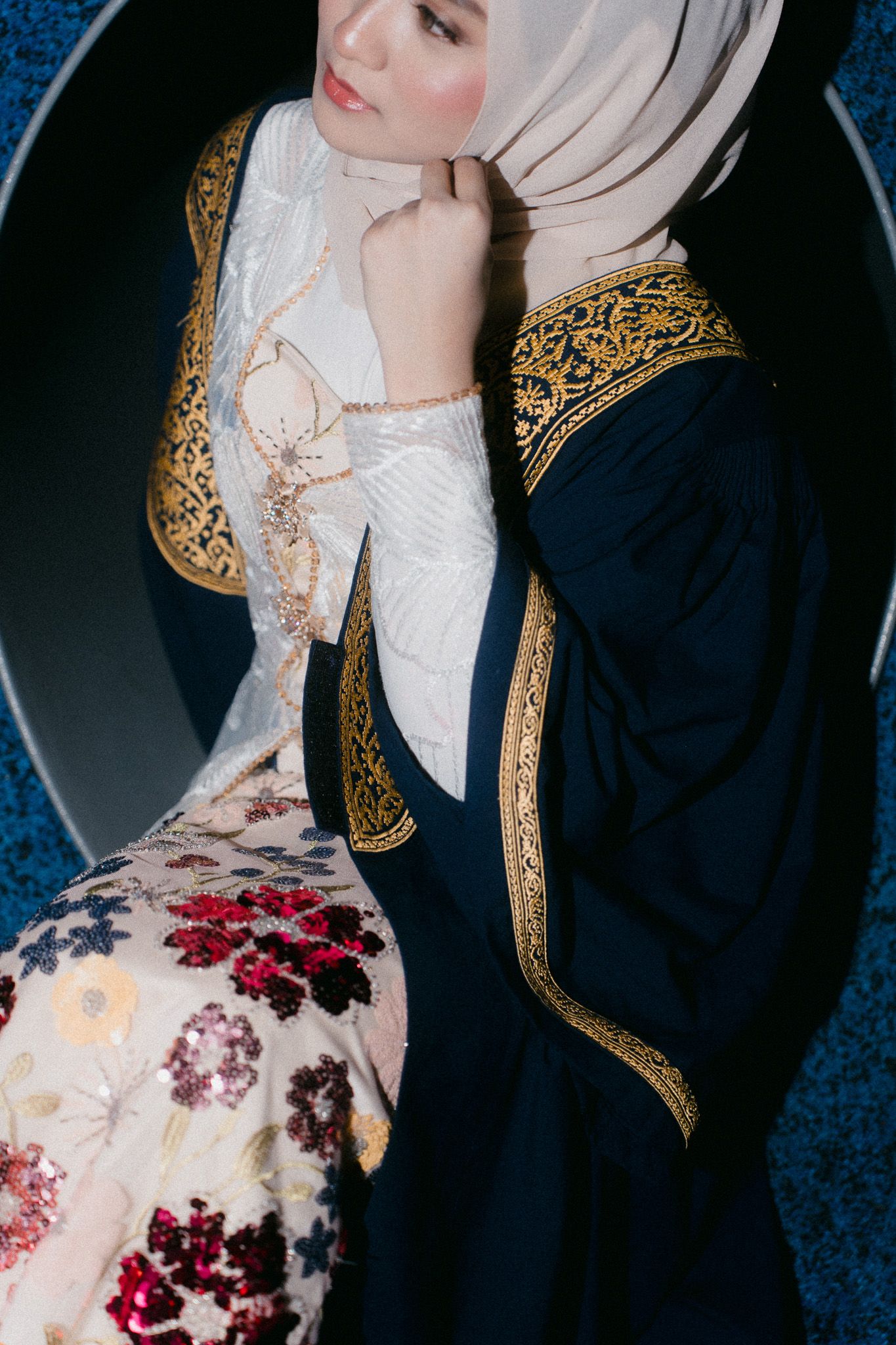 Woman in navy blue abaya with gold trim and cream hijab adjusting her headscarf near a mirror.