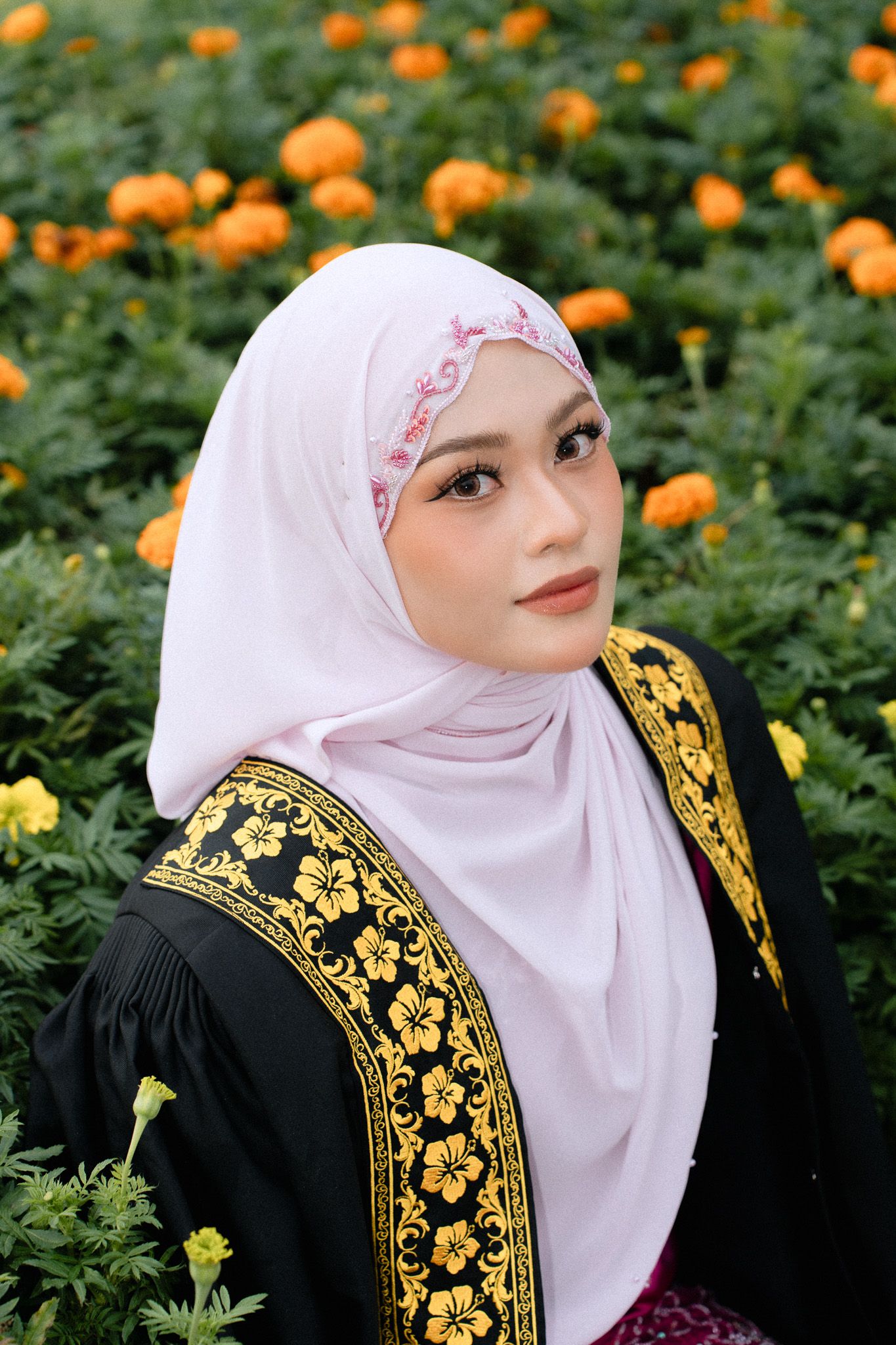 Young Muslim woman in white hijab and black embroidered dress poses gracefully among vibrant orange marigold flowers.