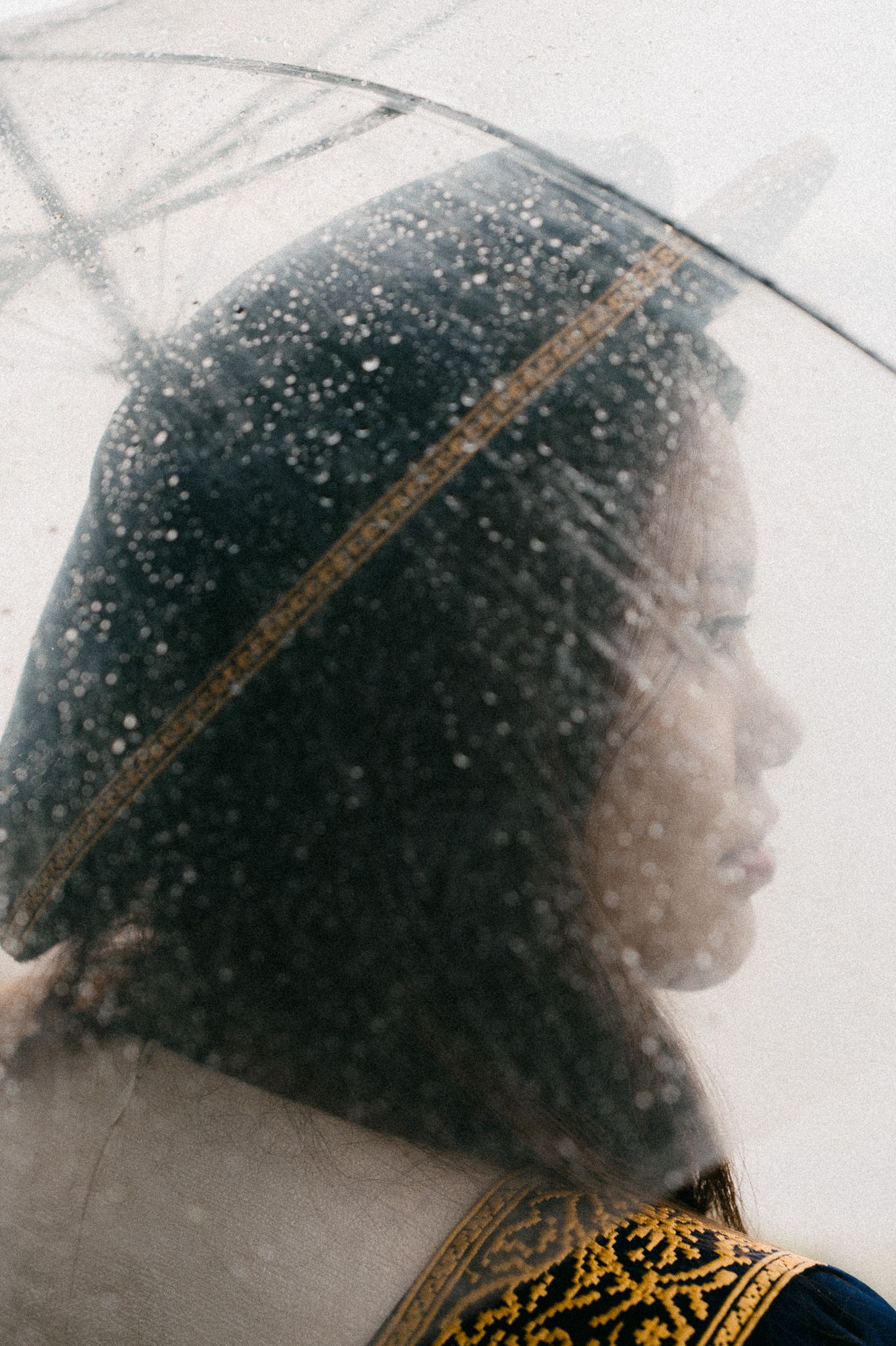 Young woman in profile under a clear umbrella with rain droplets, wearing a dark beret and embroidered top.
