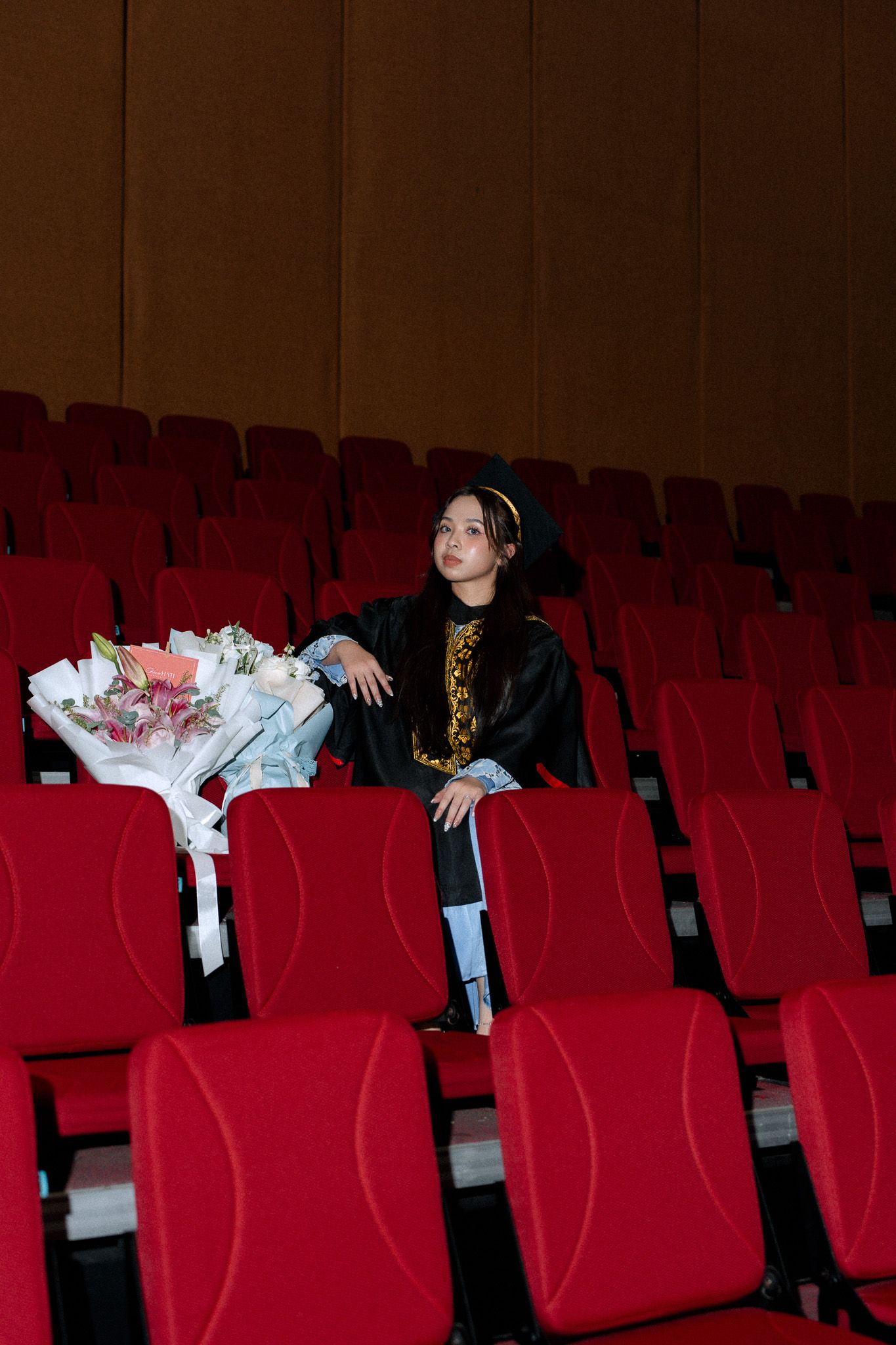Graduate in cap and gown sits alone in red theater seats holding a bouquet of flowers.