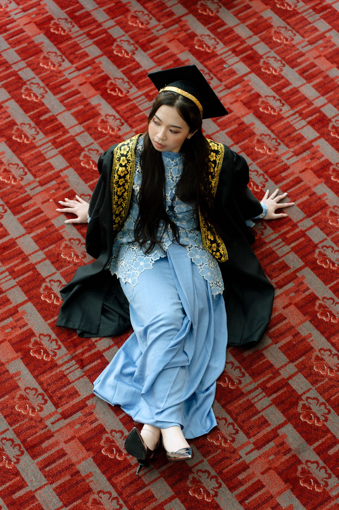 Young woman in graduation gown and cap sitting on red patterned carpet, wearing a light blue dress underneath.
