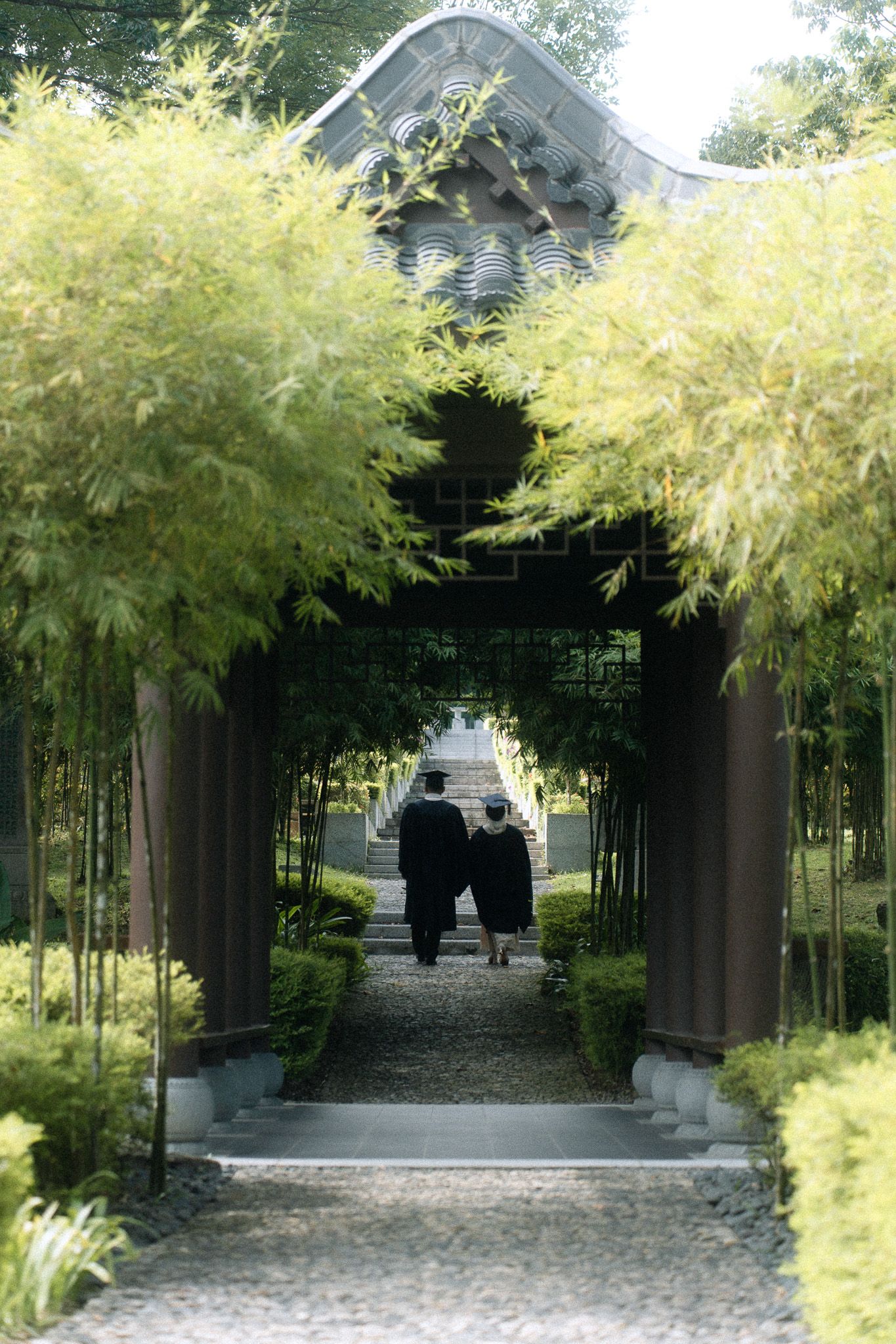 Two people in dark robes walk through a bamboo-lined garden path toward a traditional Asian temple gate.