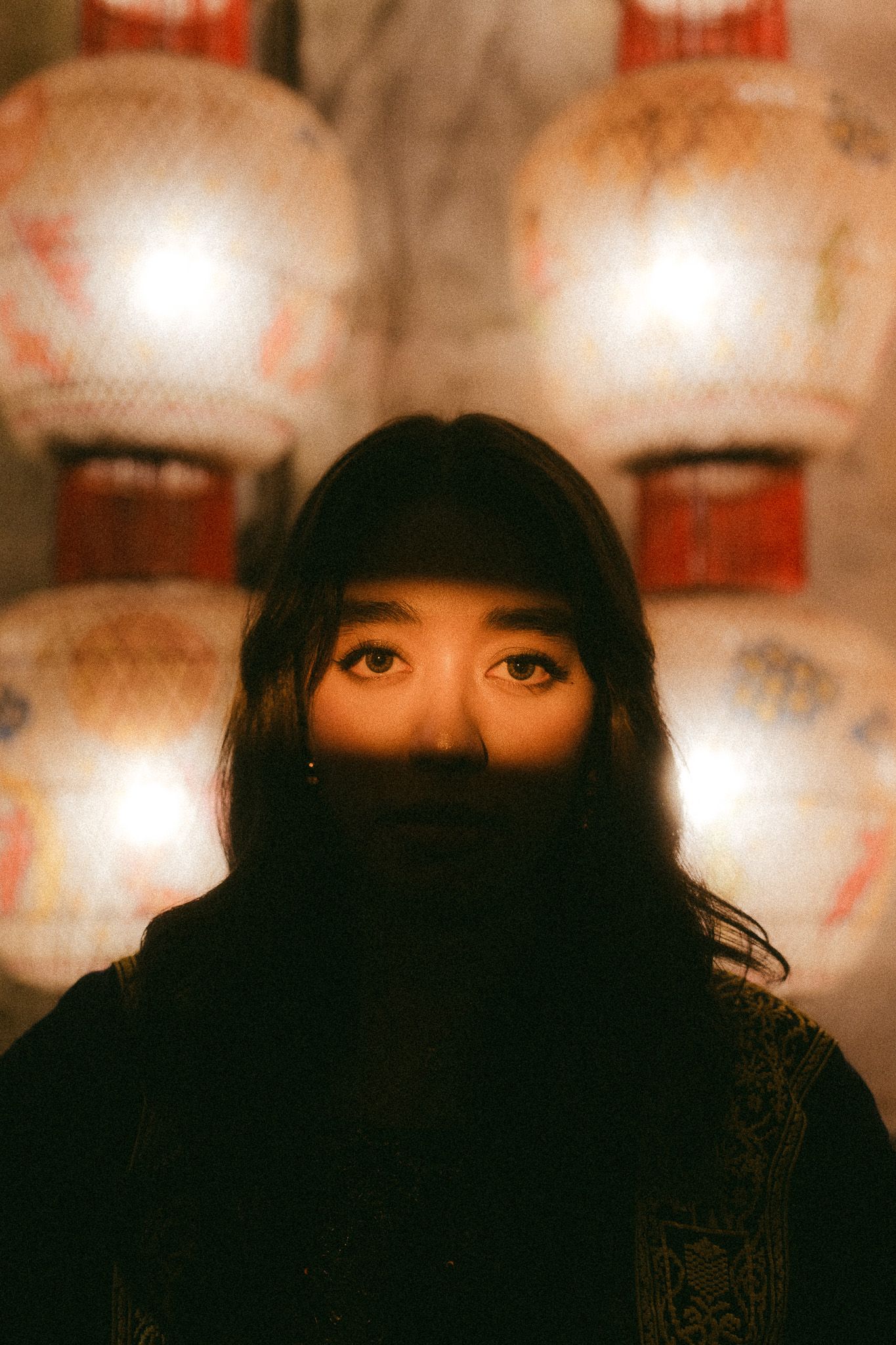 A young Asian woman with dark hair stands before glowing traditional paper lanterns in a moody, atmospheric portrait.