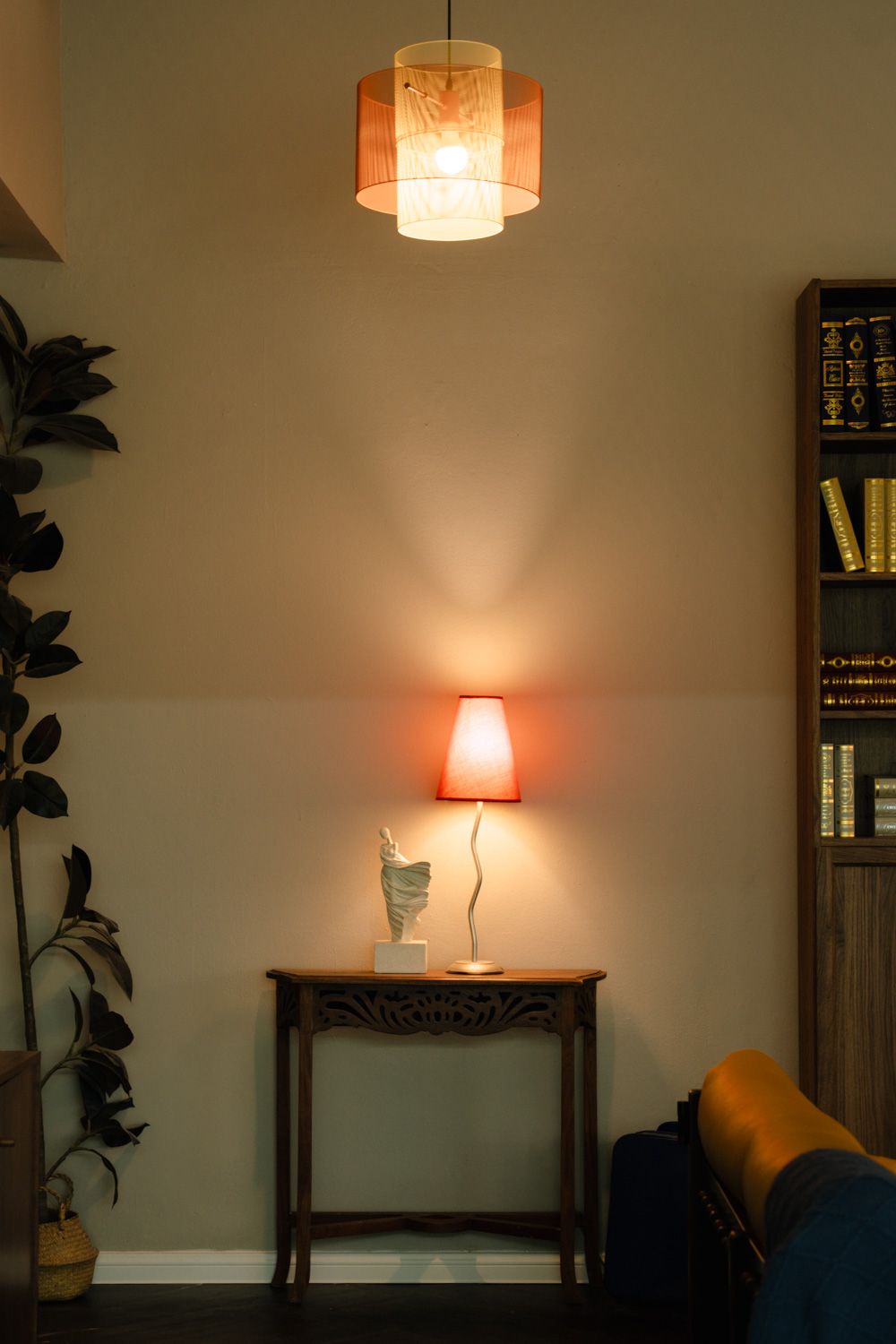 Cozy living room corner with a glowing pendant light, red table lamp on wooden console, bookshelf, and lush plant.