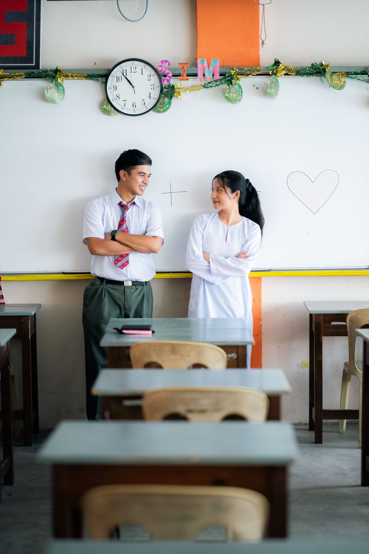 Students in school uniforms standing together in an empty classroom next to a whiteboard with a clock on the wall.
