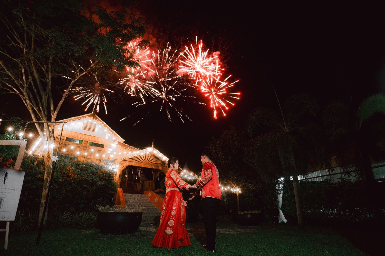 Glowing red fireworks burst against a dark night sky over illuminated outdoor structures.