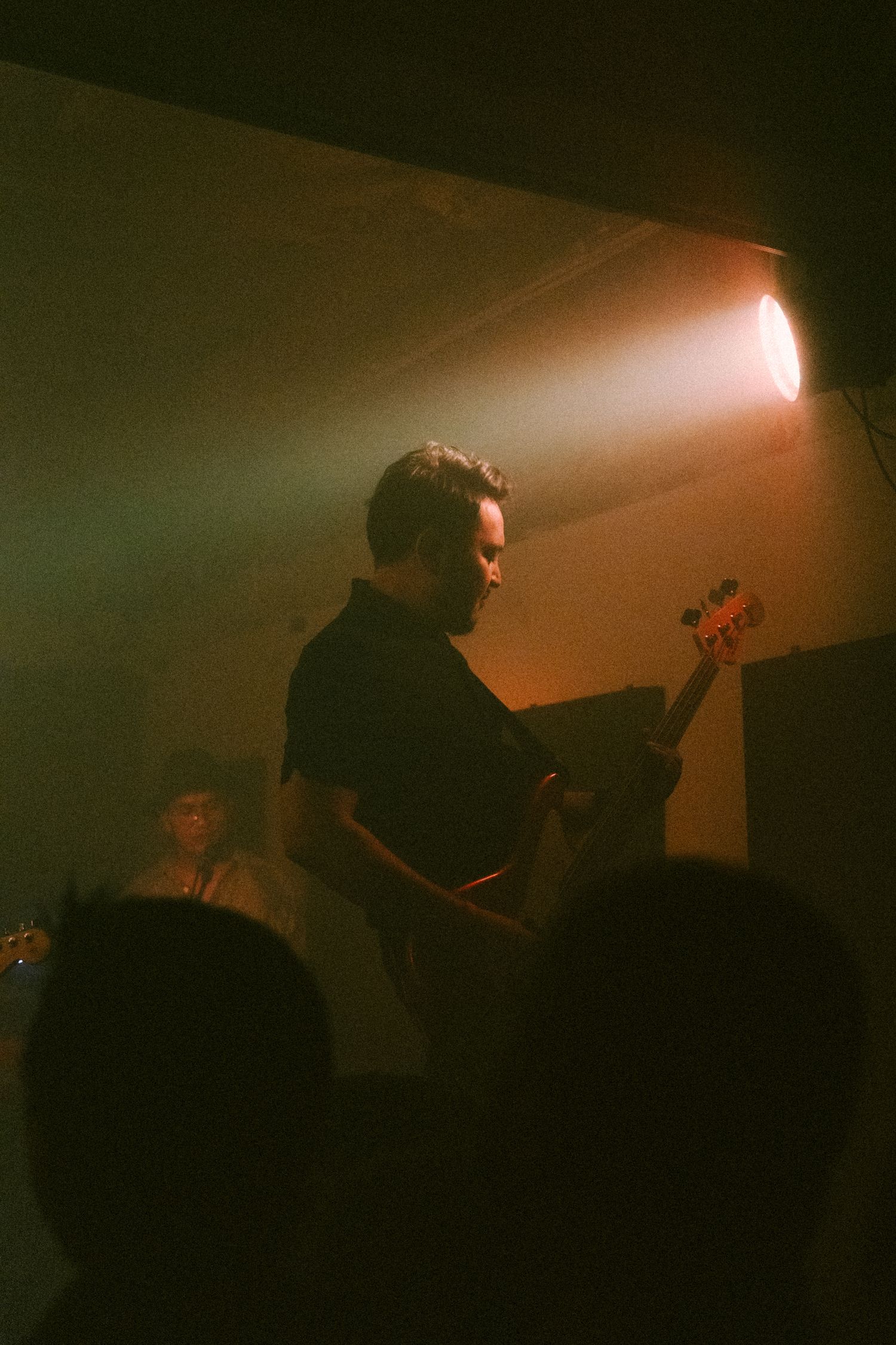 A bassist performs on stage in moody lighting with an orange spotlight beam cutting through atmospheric haze.