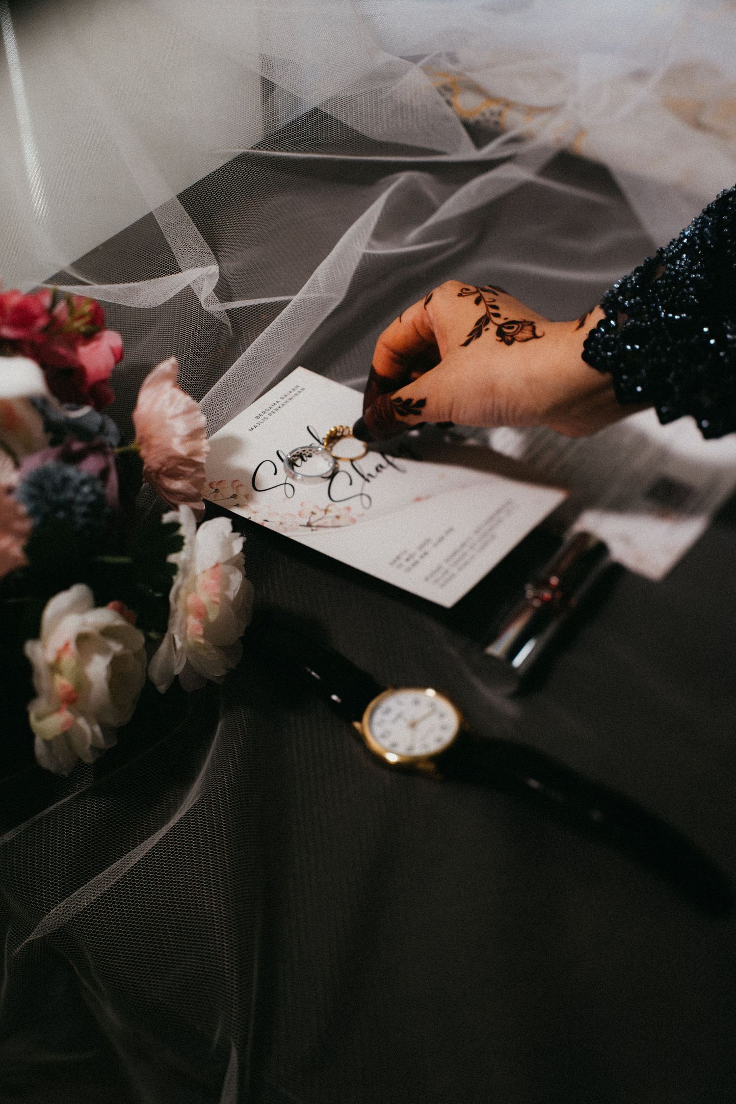 A hand signing a document with tulle fabric, flowers, and a wristwatch on a dark surface in moody lighting.