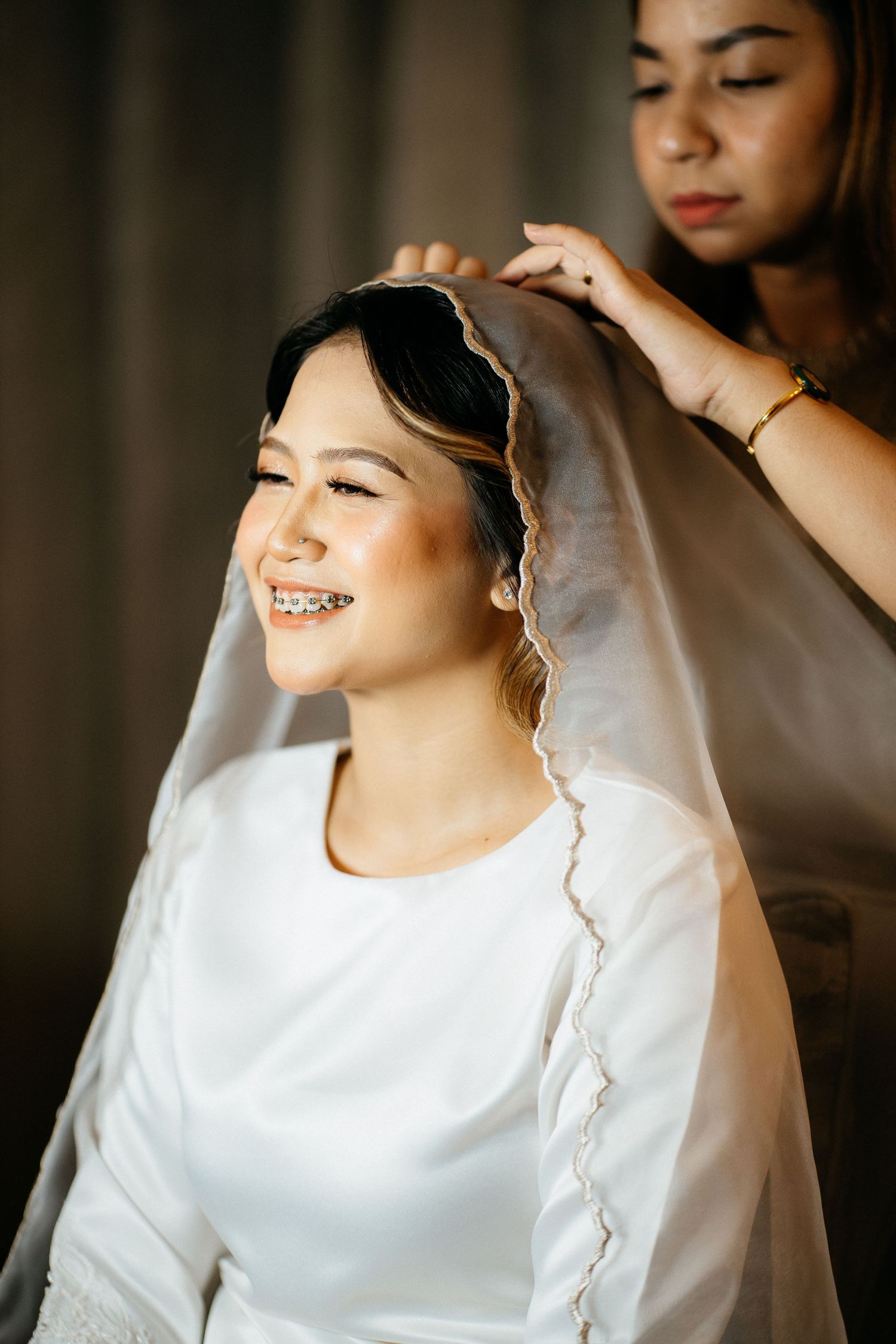A bride in a white dress sits while getting her makeup professionally done in warm lighting.