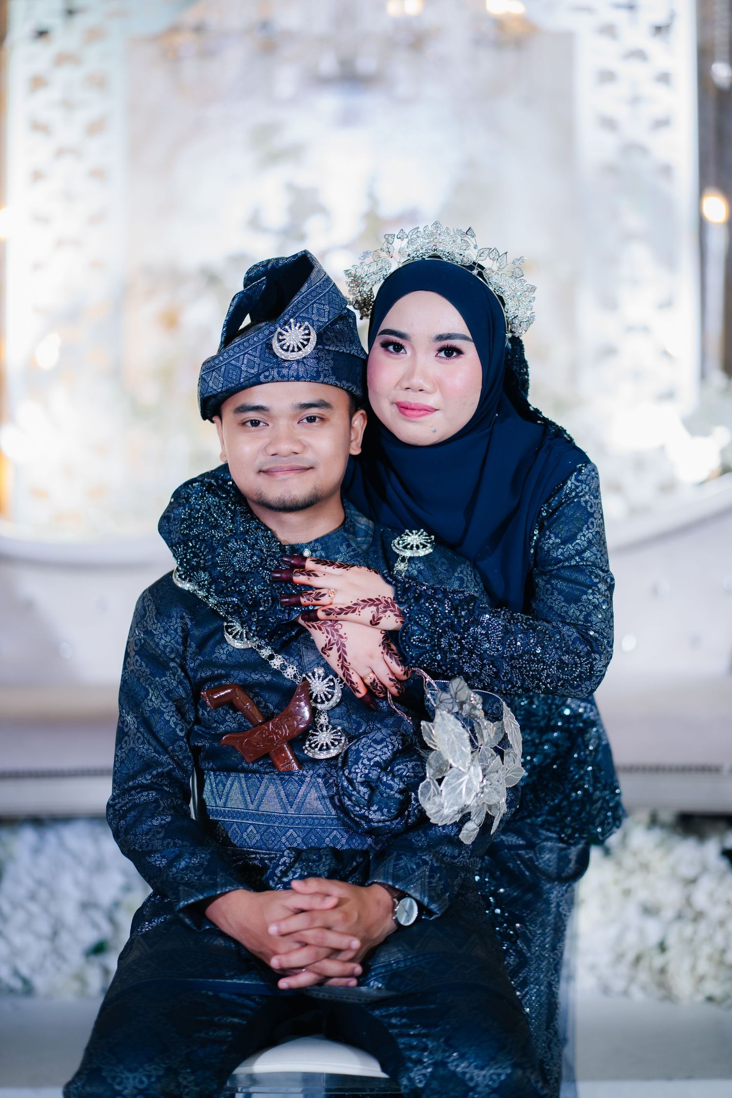 A couple in traditional Malay wedding attire with navy blue outfits and formal accessories pose together for a portrait.
