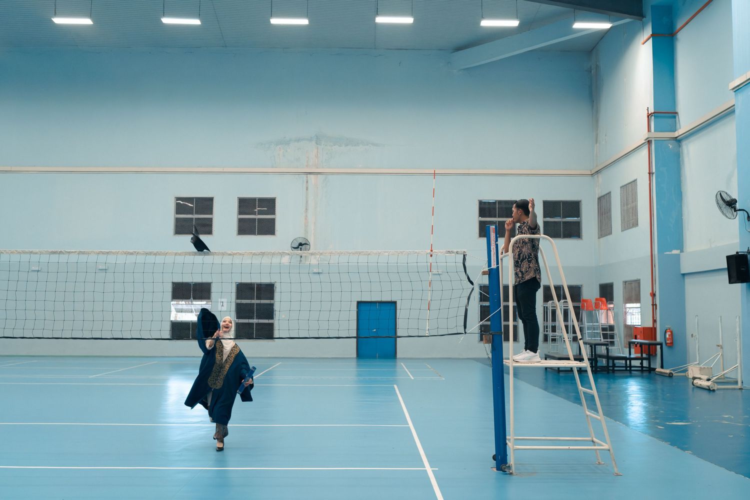 Students playing volleyball in a school gymnasium with blue flooring and white net.