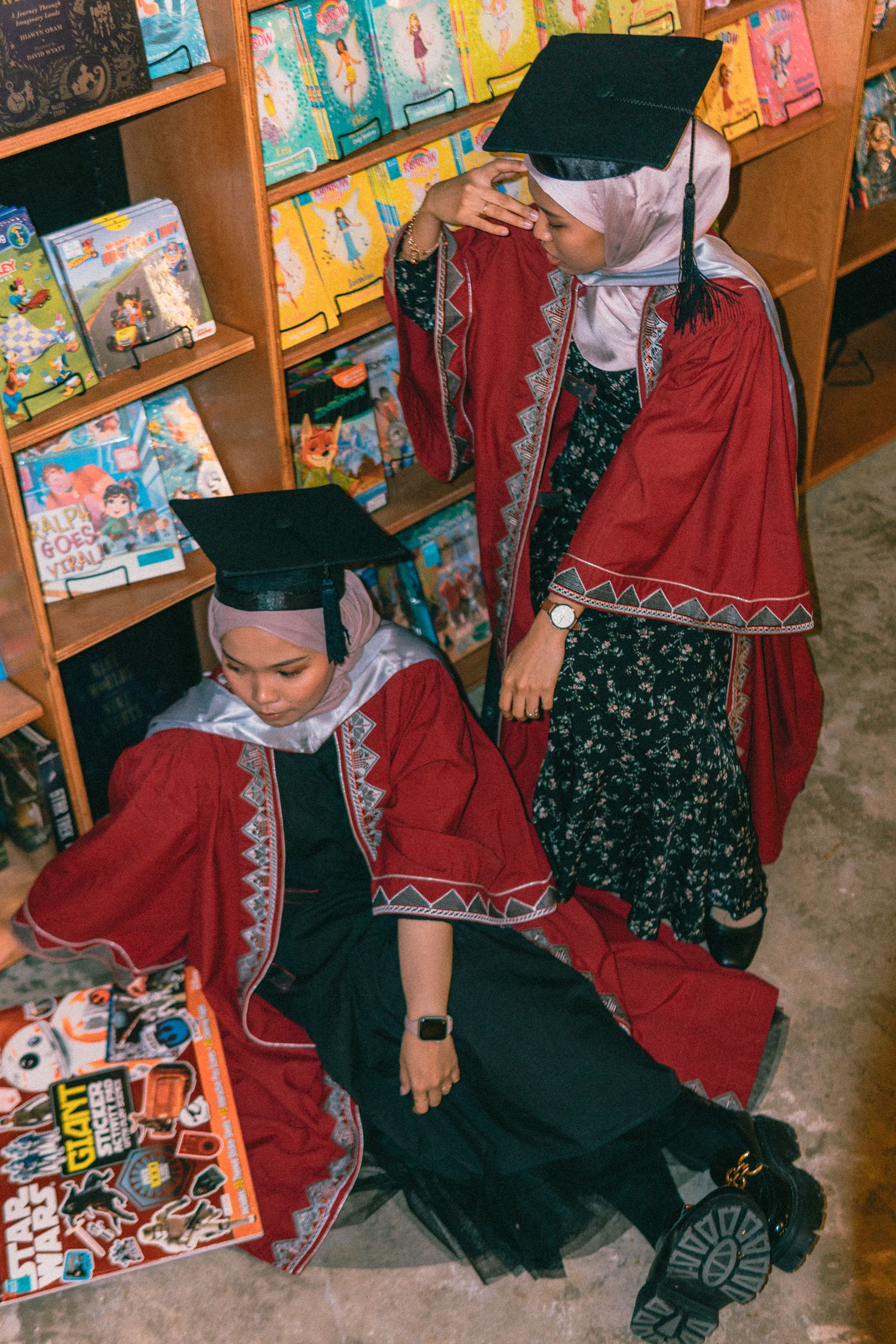 Graduates in red and black academic regalia pose playfully among library bookshelves.