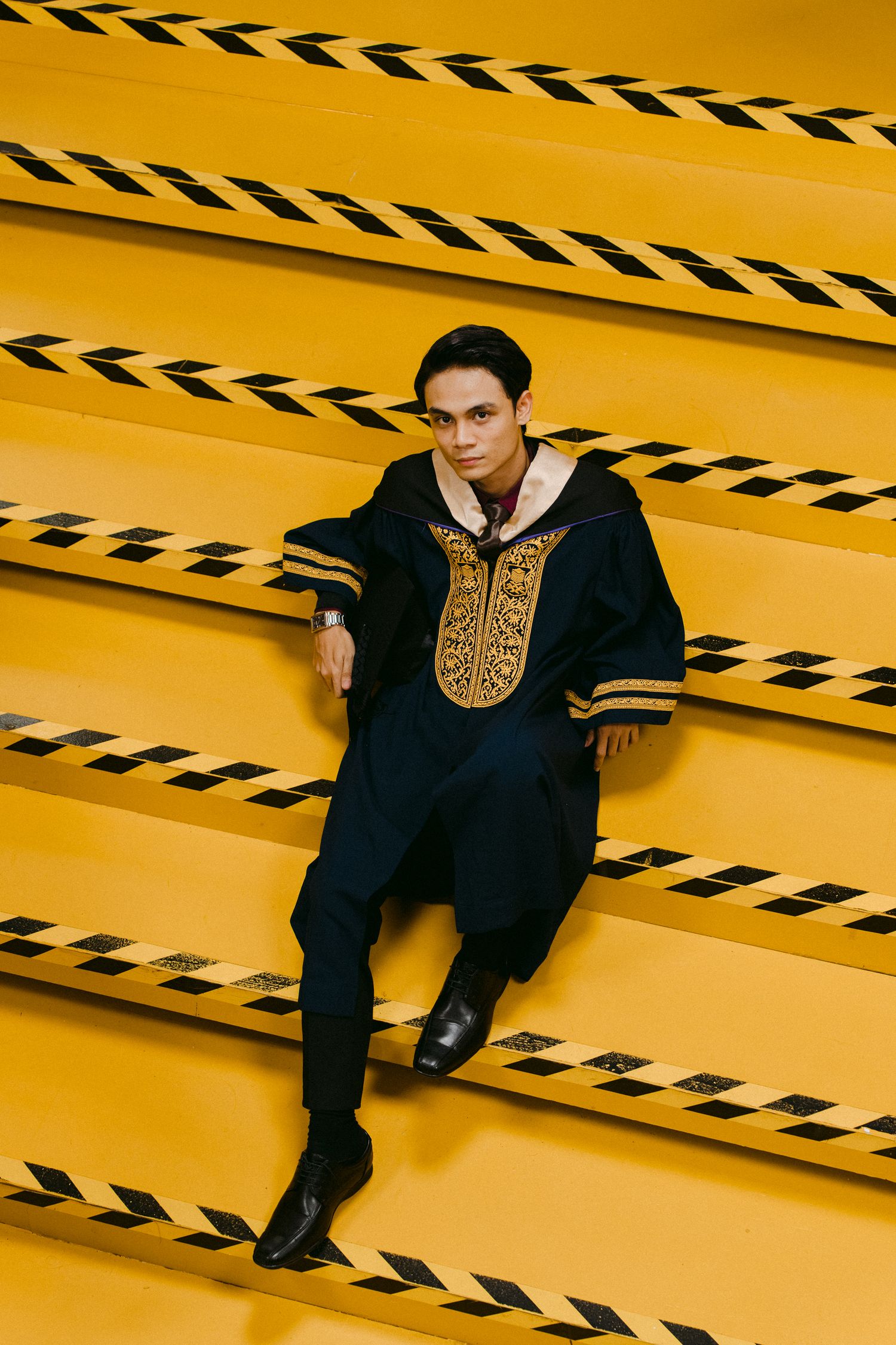 Graduate in formal black and gold attire posing on yellow steps with geometric patterns.