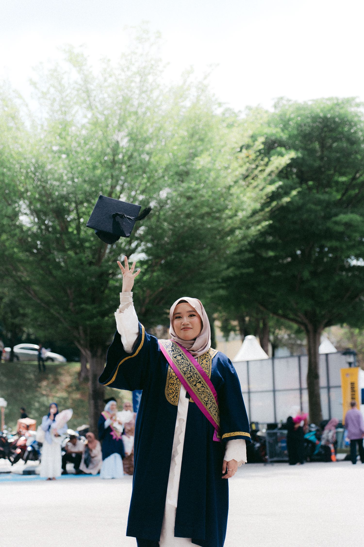 A graduate in academic regalia and hijab celebrates commencement by raising their diploma case outside on a sunny day.