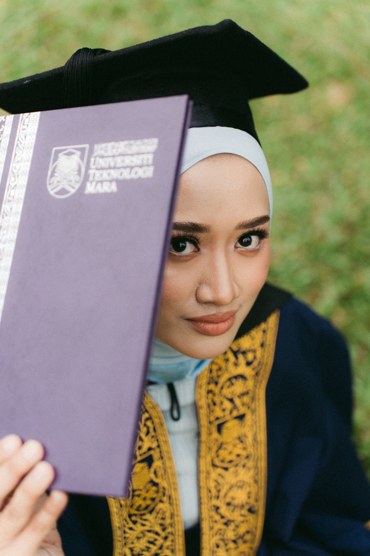 Graduate in blue hijab and academic regalia holds purple diploma folder during outdoor graduation ceremony.
