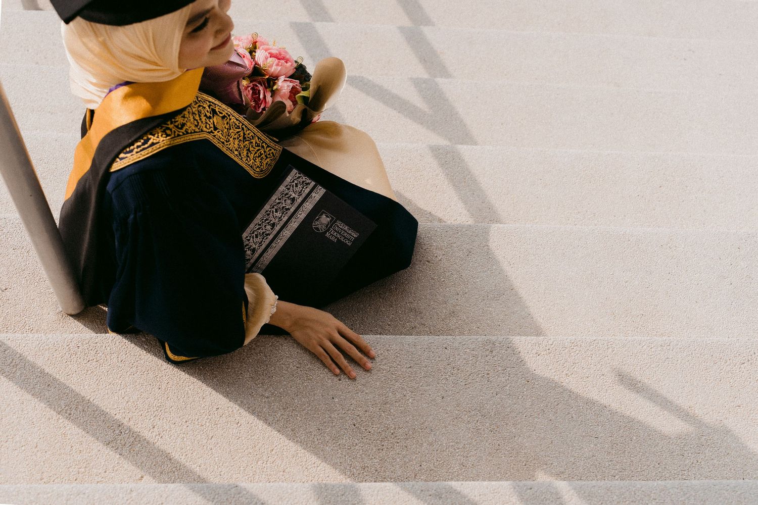 Person in traditional black and gold embroidered garment praying on floor while casting shadows across light surface.