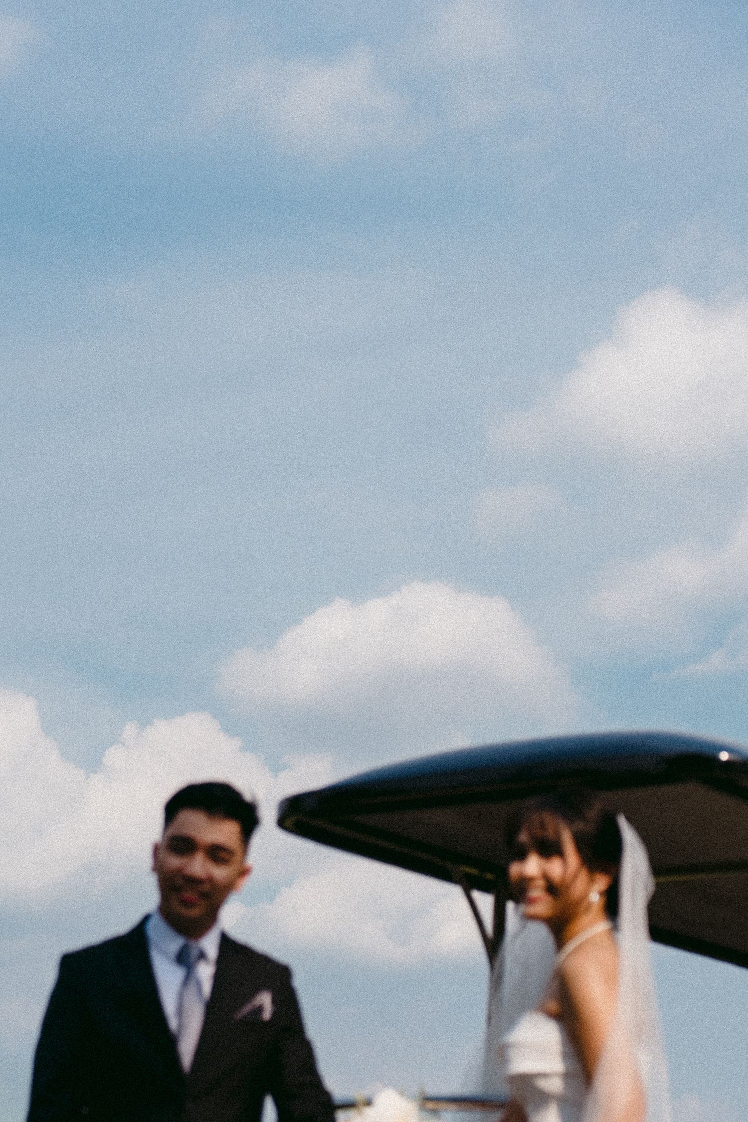 A couple in wedding attire stands together under a canopy against a blue sky with white clouds.