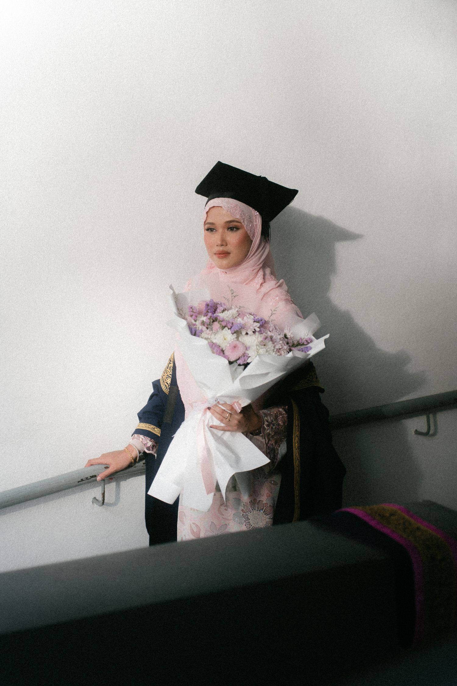A figure in a pink hijab and graduation cap poses dramatically against a white wall in a triptych series of photos.