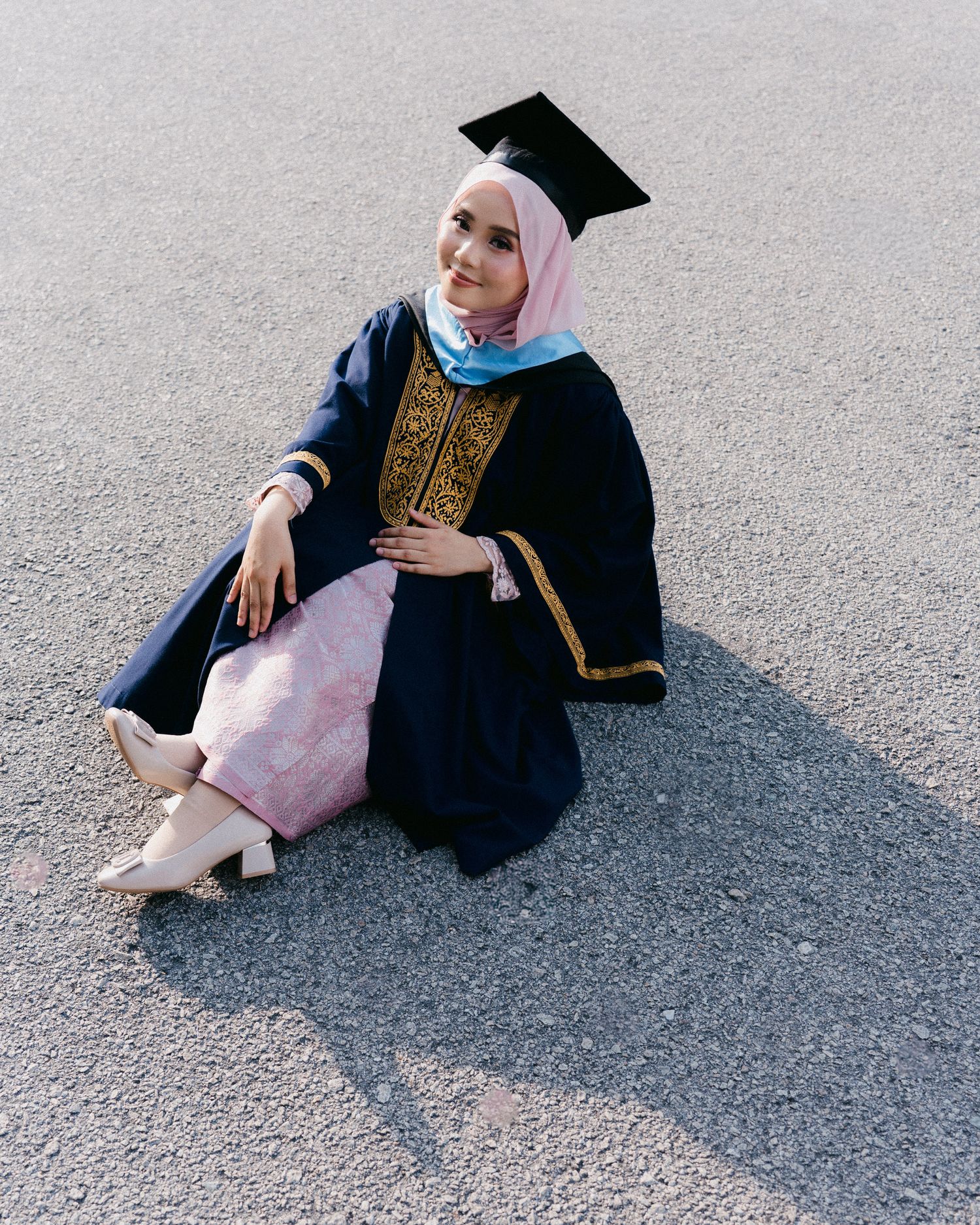 Graduate in navy academic regalia with gold embroidery and pink hijab sits on ground during graduation photoshoot.
