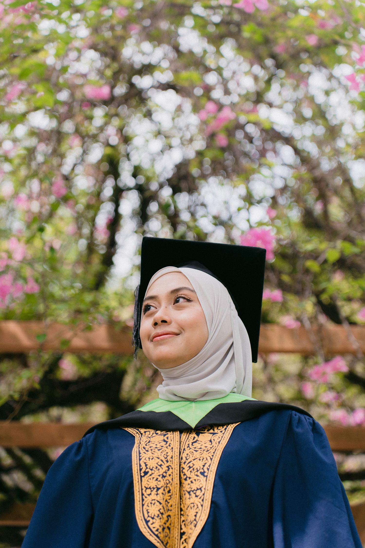 Graduate in blue gown and white hijab poses in front of blooming cherry blossoms during spring graduation ceremony.