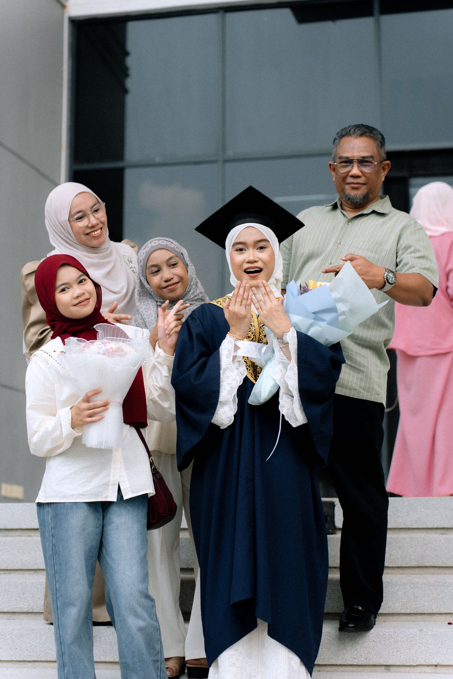 A group of graduates in hijabs and casual wear celebrate together on outdoor steps while holding papers and laughing.