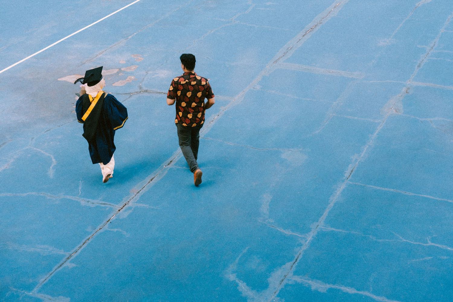 Two people walking on a bright blue surface, viewed from above, with one wearing a graduation gown.