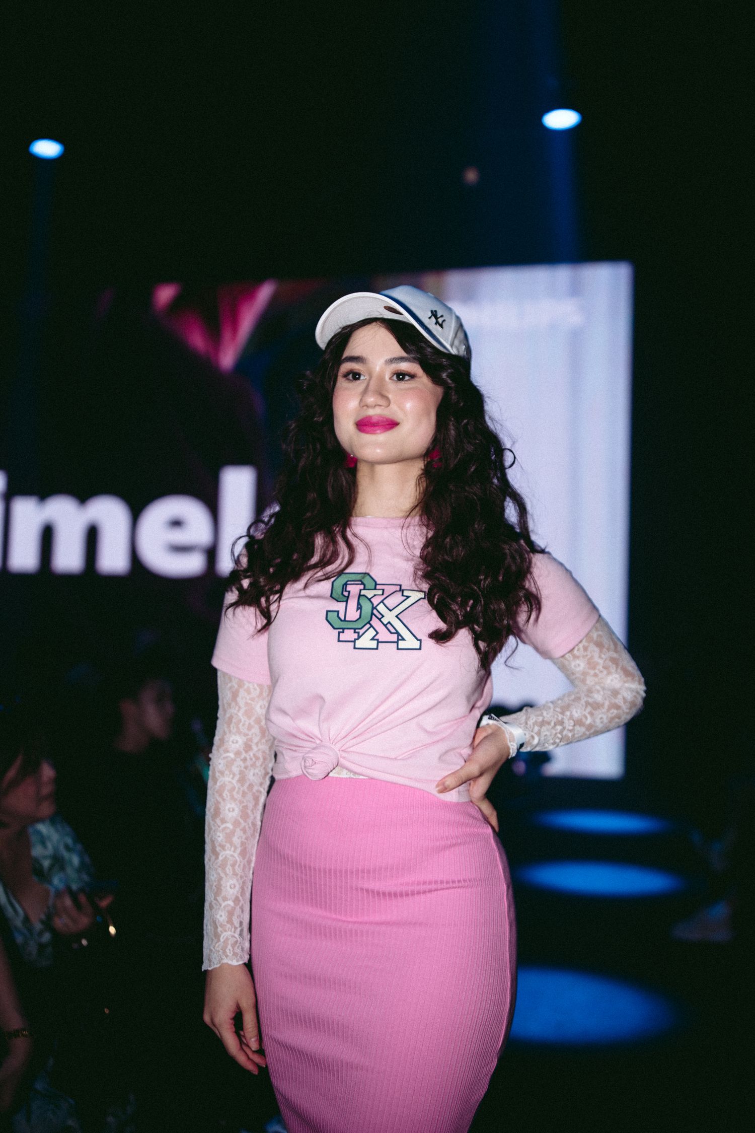 Model in pink outfit and baseball cap poses under stage lighting at a fashion event.