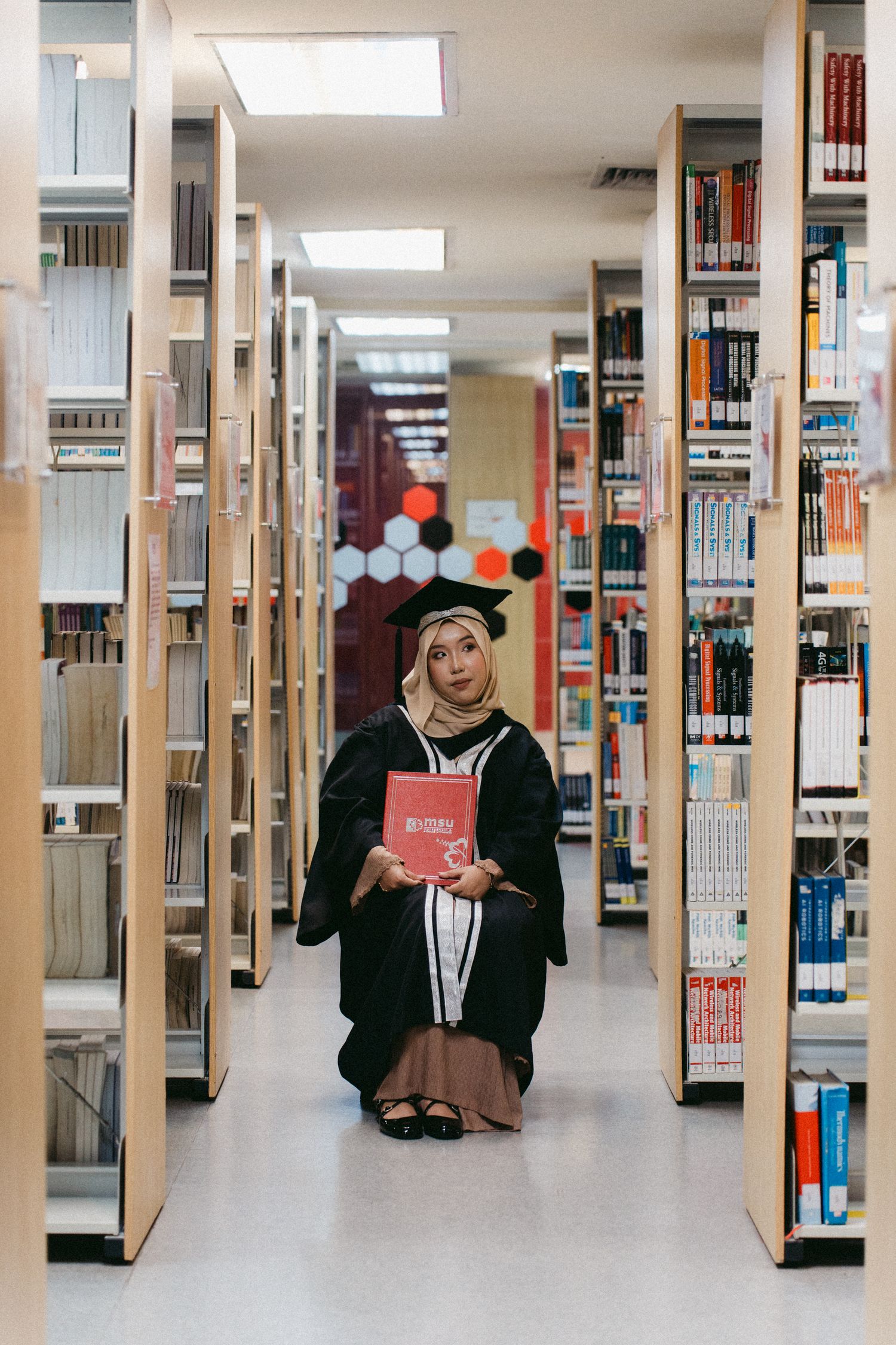 A student in a hijab sits on the floor between library shelving while holding a red book.