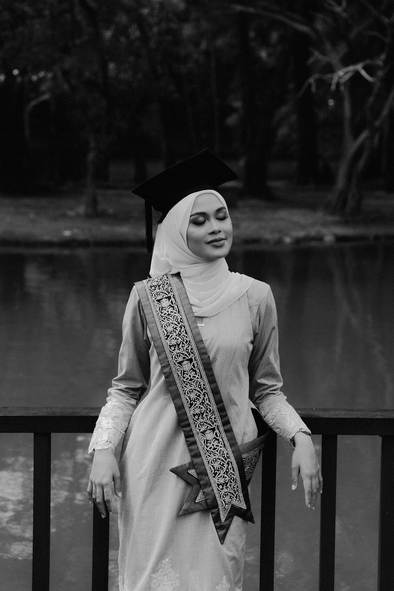 Smiling Muslim woman in hijab and graduation cap wearing a decorative sash, posing by a lake in black and white.