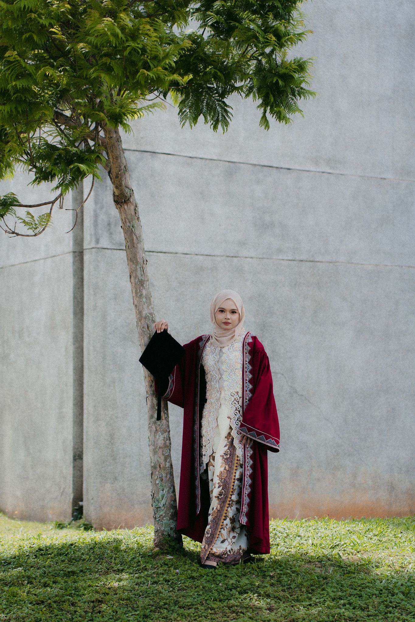 Muslim woman in hijab and maroon abaya holds graduation cap beside tree, celebrating academic achievement outdoors.