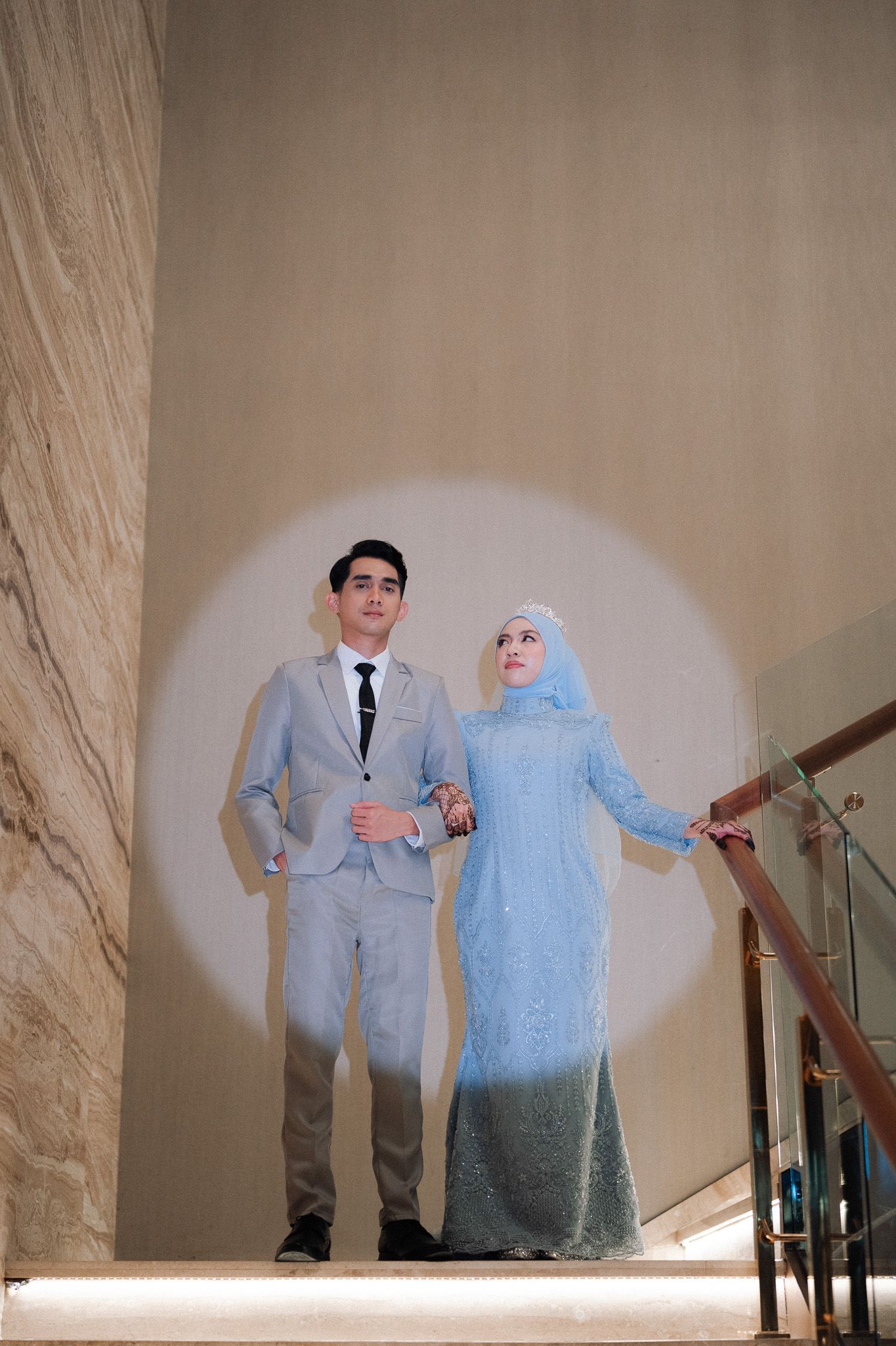 Muslim couple in matching light blue outfits posing on stairs, woman wearing hijab, man in suit with black tie.